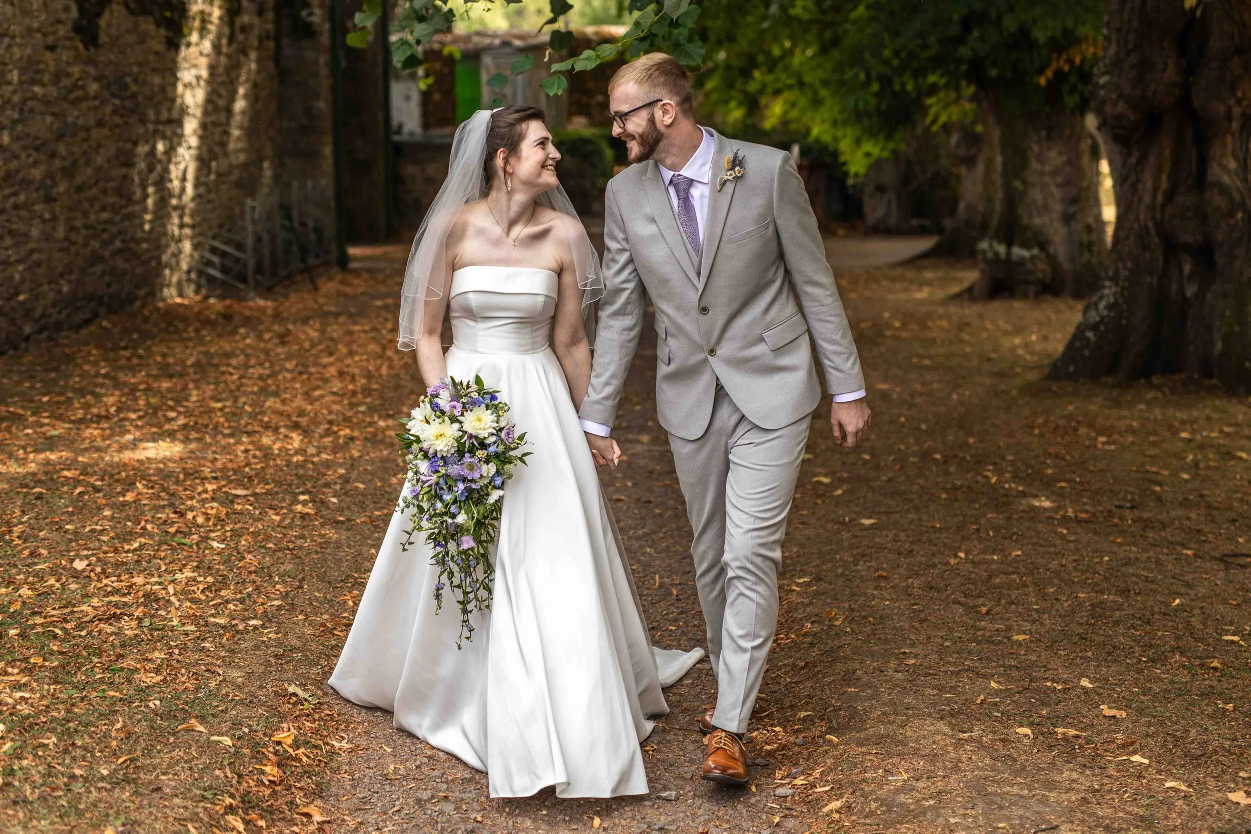 A bride and groom walk hand in hand through a park covered with fallen autumn leaves. The bride wears a strapless white wedding dress and a veil, and holds a bouquet of purple and white flowers. The groom wears a light gray suit with a purple tie and