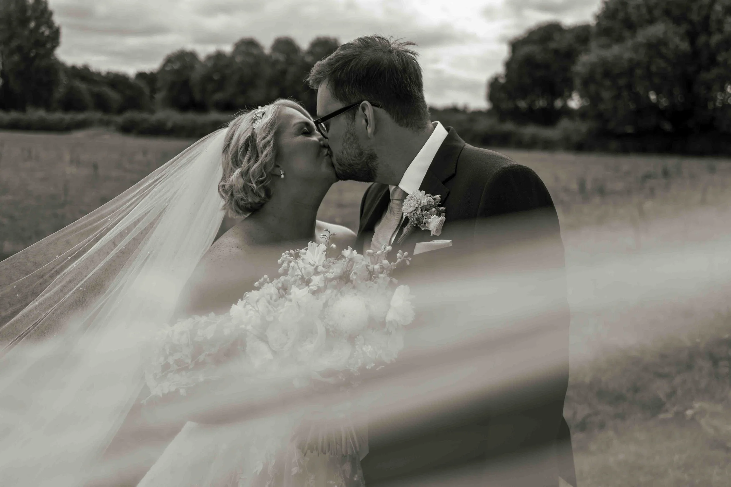 A black and white photo of a bride and groom kissing outdoors on a cloudy day, with trees in the background. The bride wears a veil and has short curly hair, while the groom wears glasses and a suit with a boutonnière.