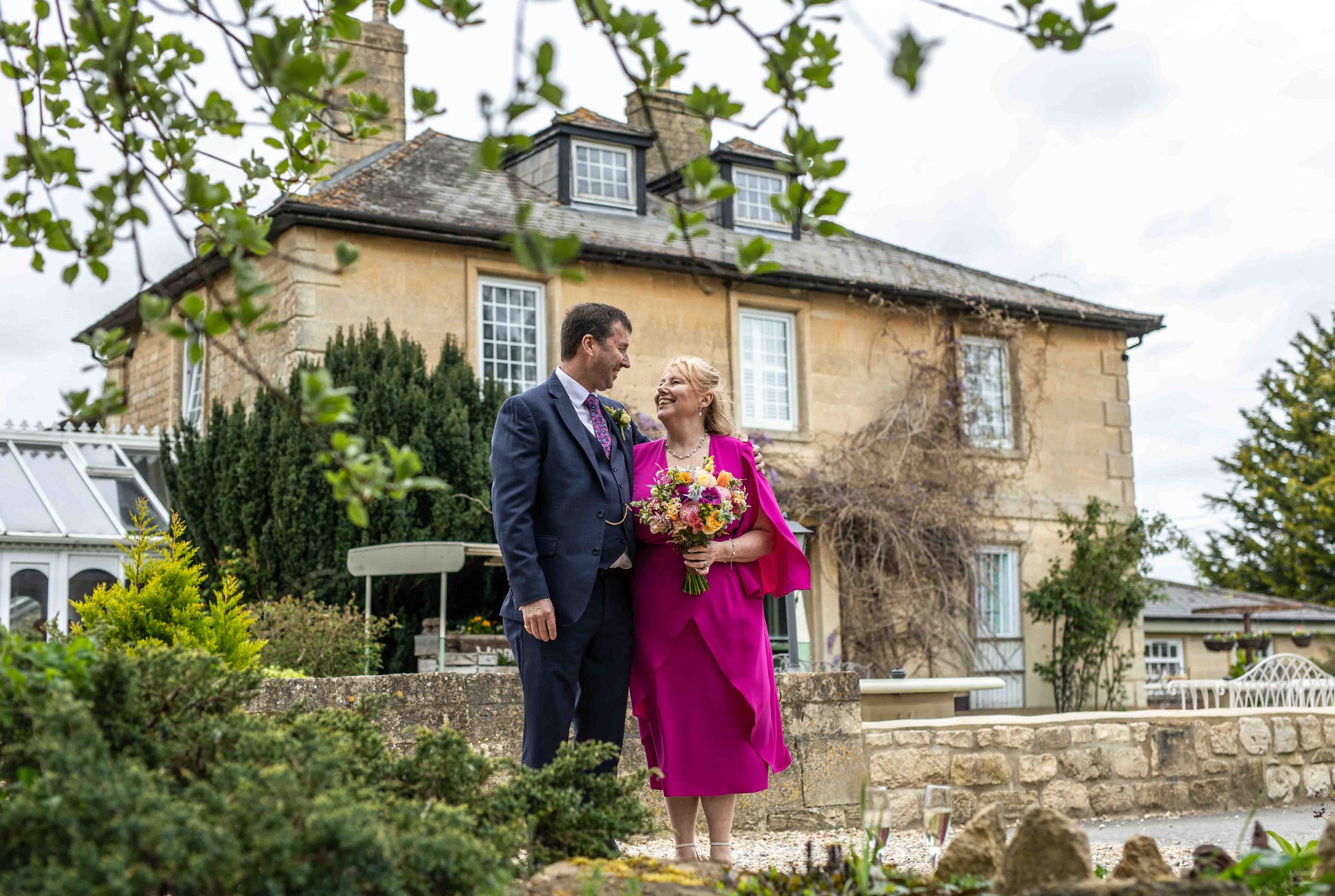 A couple, a man and a woman, stand close together outdoors in front of a large house, smiling at each other. The woman is holding a bouquet of colorful flowers and wearing a bright pink dress, and the man is dressed in a dark suit with a floral tie. 