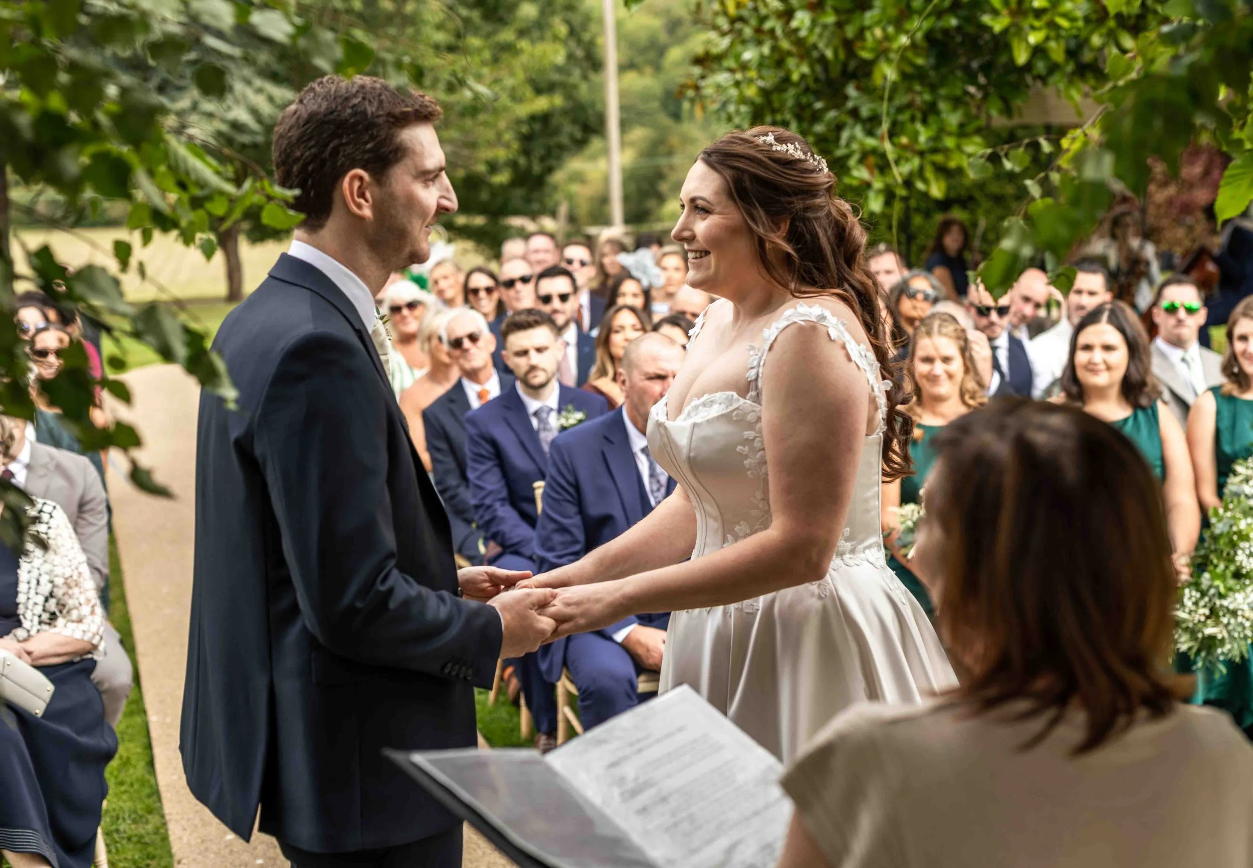 A bride and groom exchange vows outdoors during a wedding ceremony with guests seated behind them, surrounded by greenery.