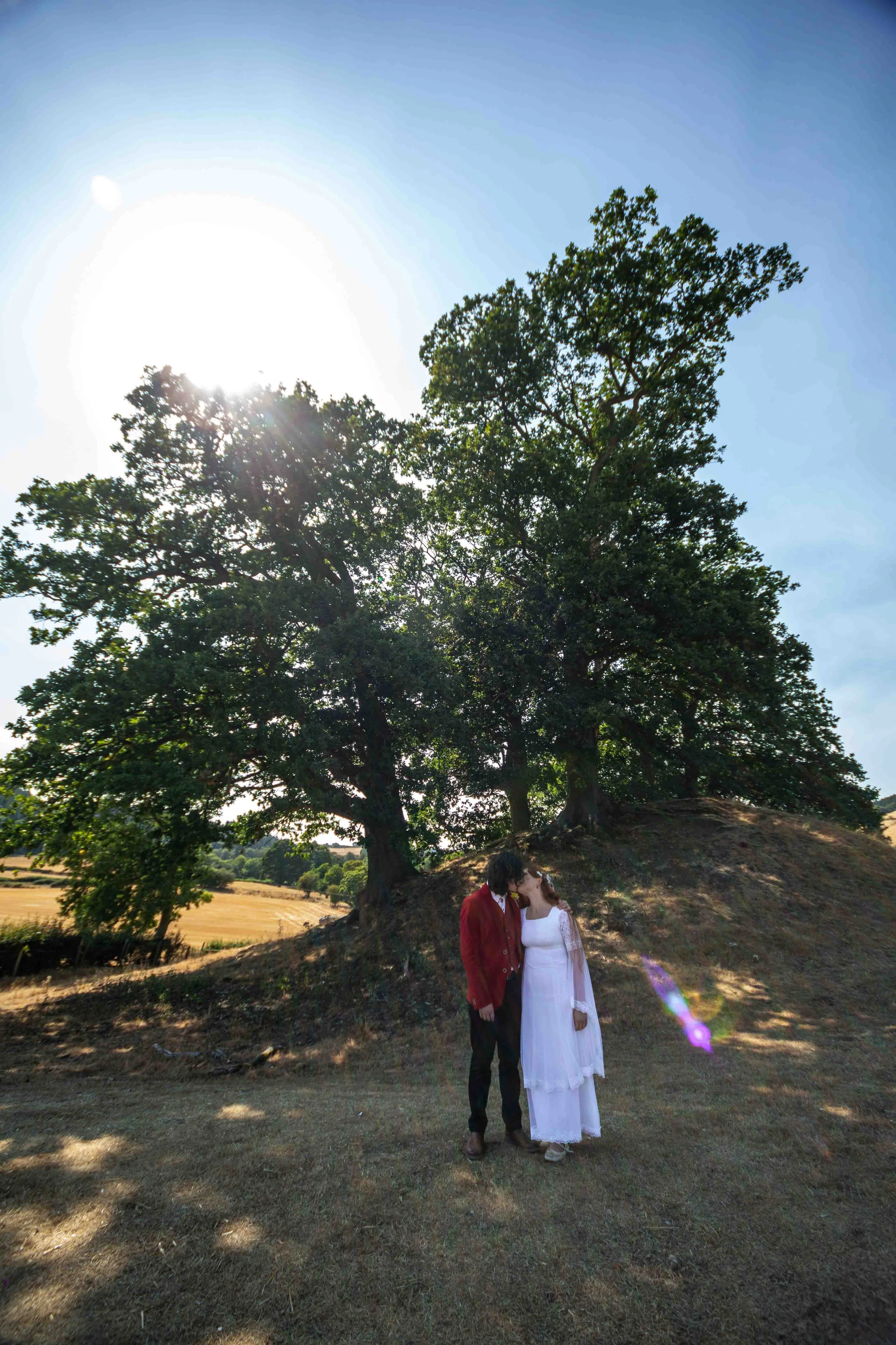 A couple shares a kiss under a large tree in a sunny open field. The man is wearing a red jacket and dark pants, and the woman is dressed in a white lace dress. The sun is shining brightly from behind the tree, creating a lens flare in the bottom rig