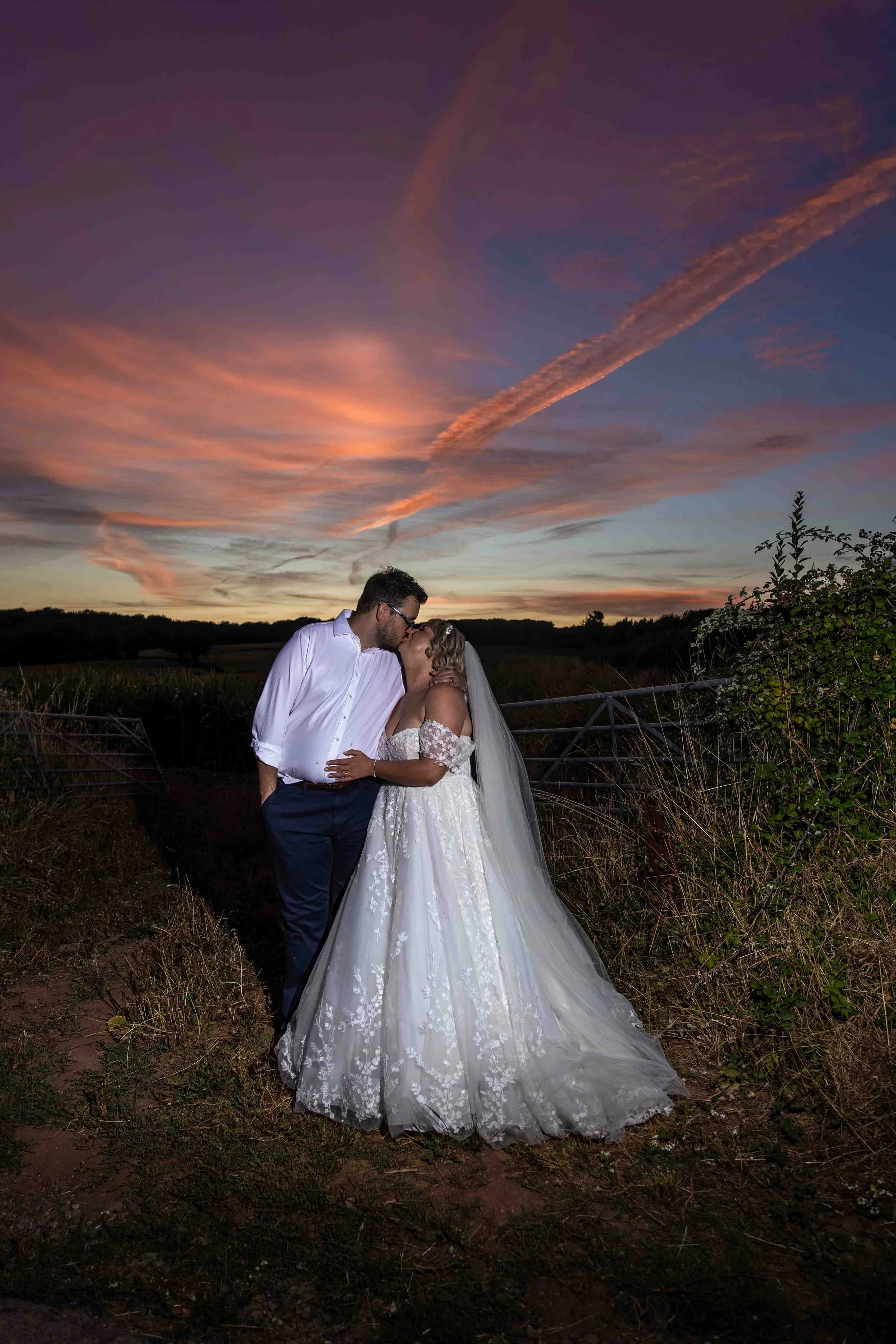 A bride and groom sharing a kiss outdoors at sunset, with a colorful sky and clouds in the background.