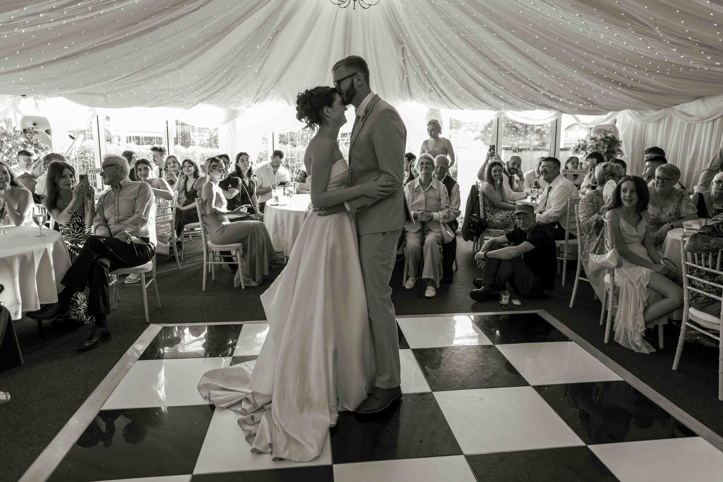 A bride and groom kissing on a dance floor at their wedding reception, with wedding guests seated at round tables, smiling and watching. The reception is decorated with draped ceiling fabric and string lights.