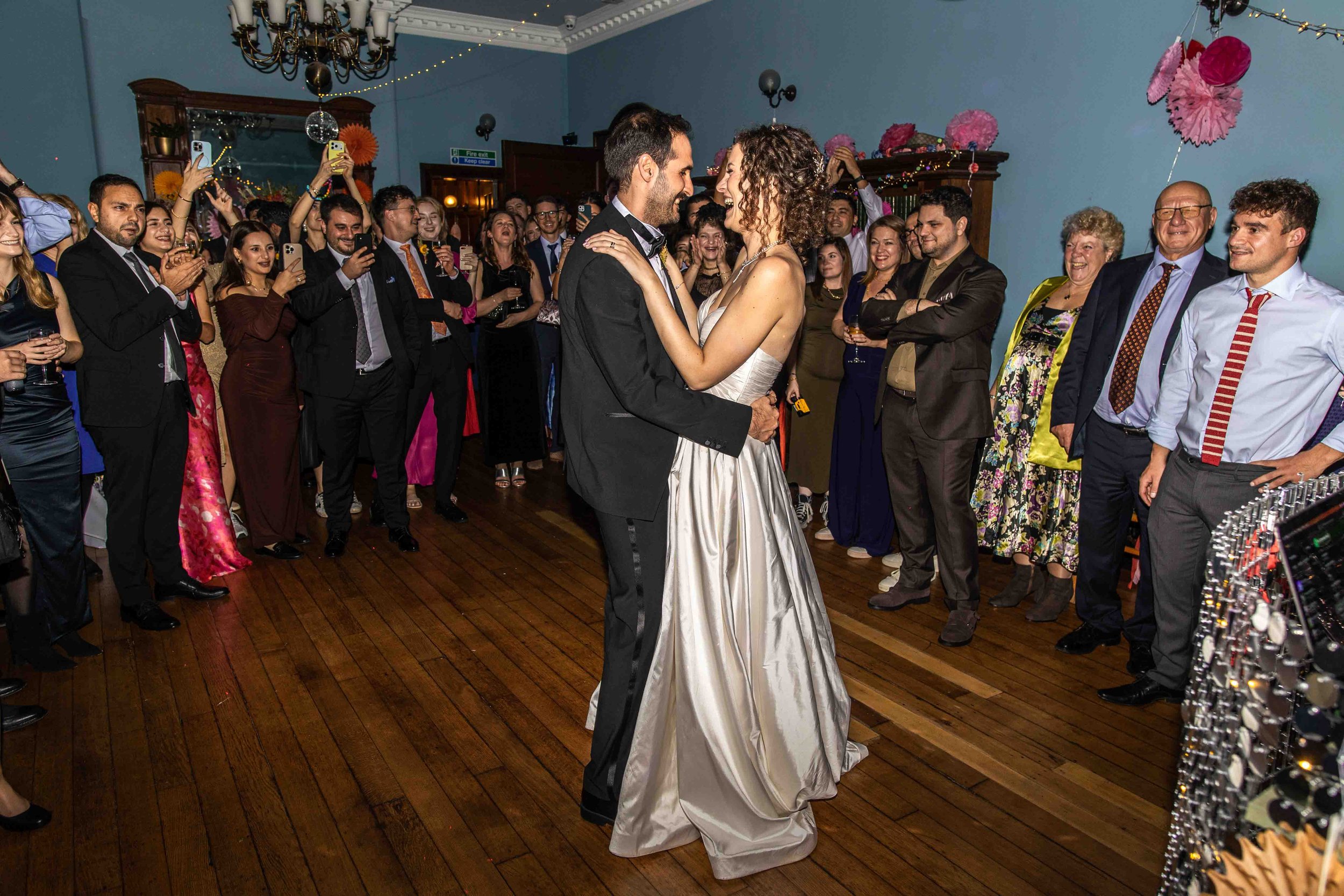 A couple in wedding attire dances closely at a wedding reception while surrounded by guests smiling, taking photos, and enjoying the celebration in a decorated indoor venue.