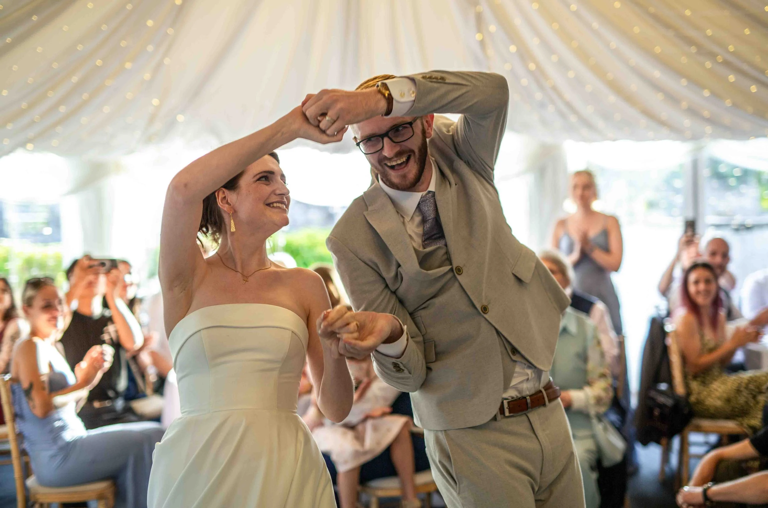 A bride and groom are dancing together at their wedding reception. The groom is wearing a light-colored suit and glasses, while the bride is dressed in a strapless white wedding gown. They are holding hands and smiling happily, with guests watching a