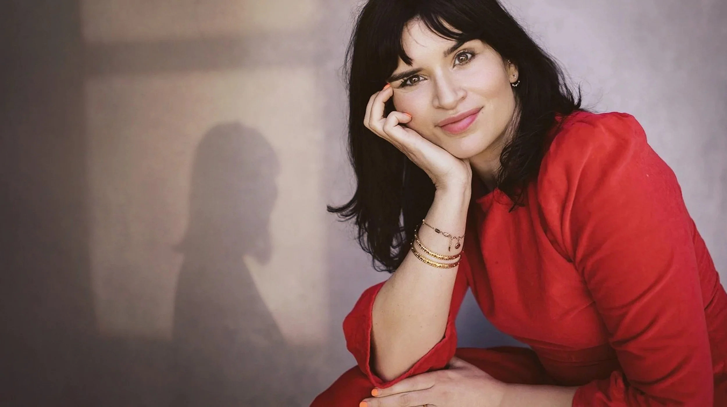 A woman with dark hair in a red top, sitting and resting her head on her hand, smiling softly, with jewelry on her wrist, against a neutral background.