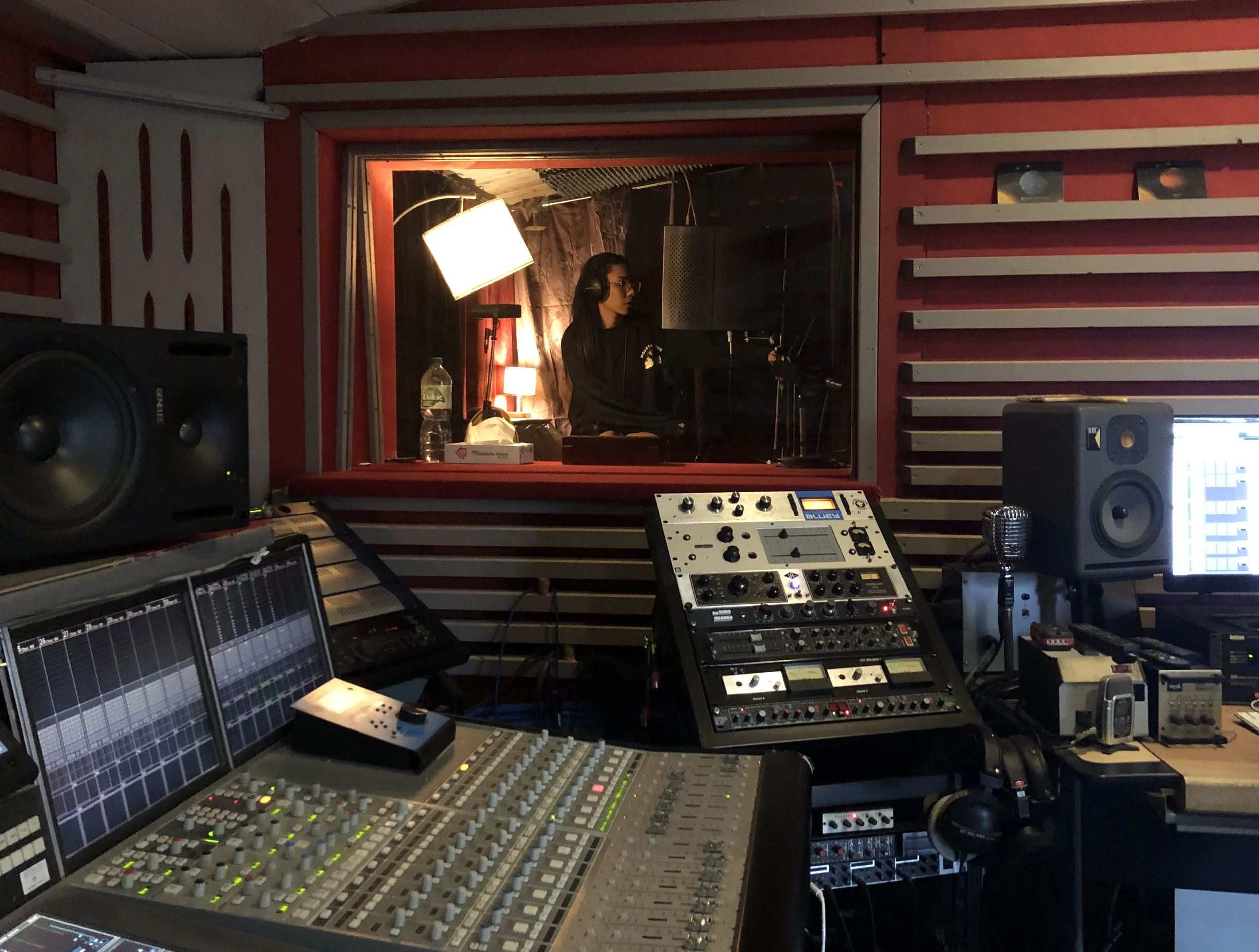 Salle d'enregistrement avec équipement audio et une jeune femme dans la cabine voix.