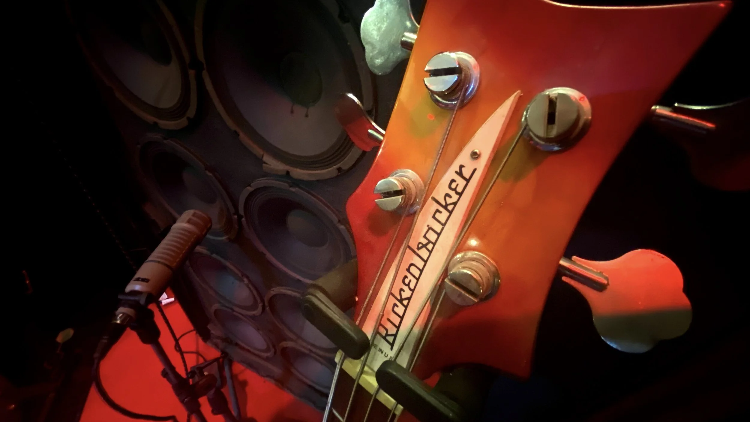 Guitare électrique orange avec marque 'Rickenbacker' visible sur la tête, micro de guitare et câbles attachés, entourée de haut-parleurs noirs.
