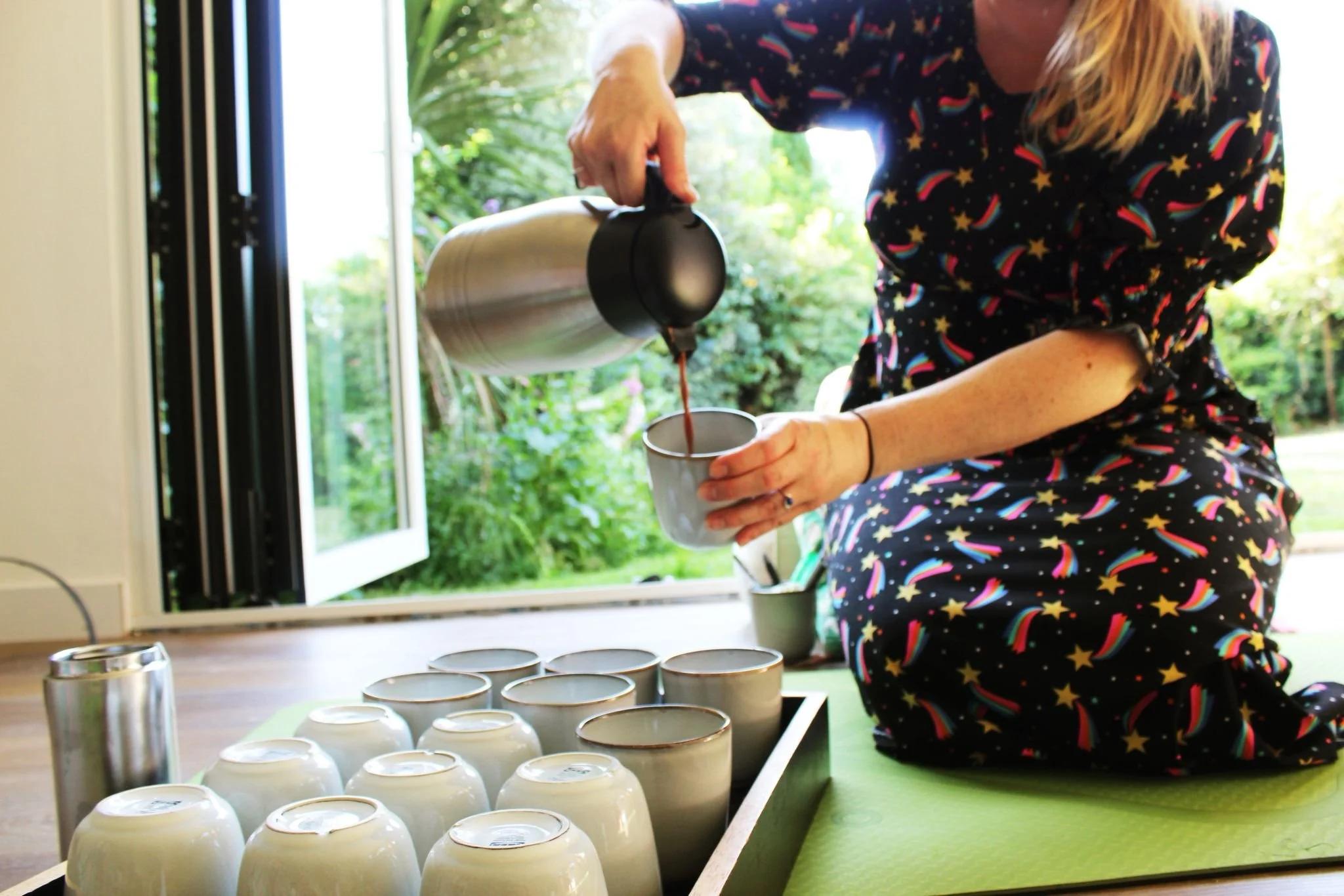 A woman pours a drink from a metallic pitcher into a white mug on a windowsill with several more mugs and a metal tumbler nearby, in a bright room with a garden view.