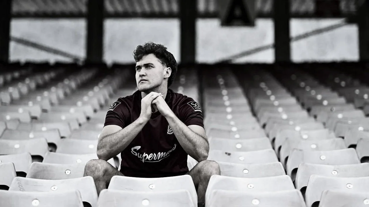 A young man sitting alone in empty stadium seats, looking contemplative with his hands clasped under his chin.