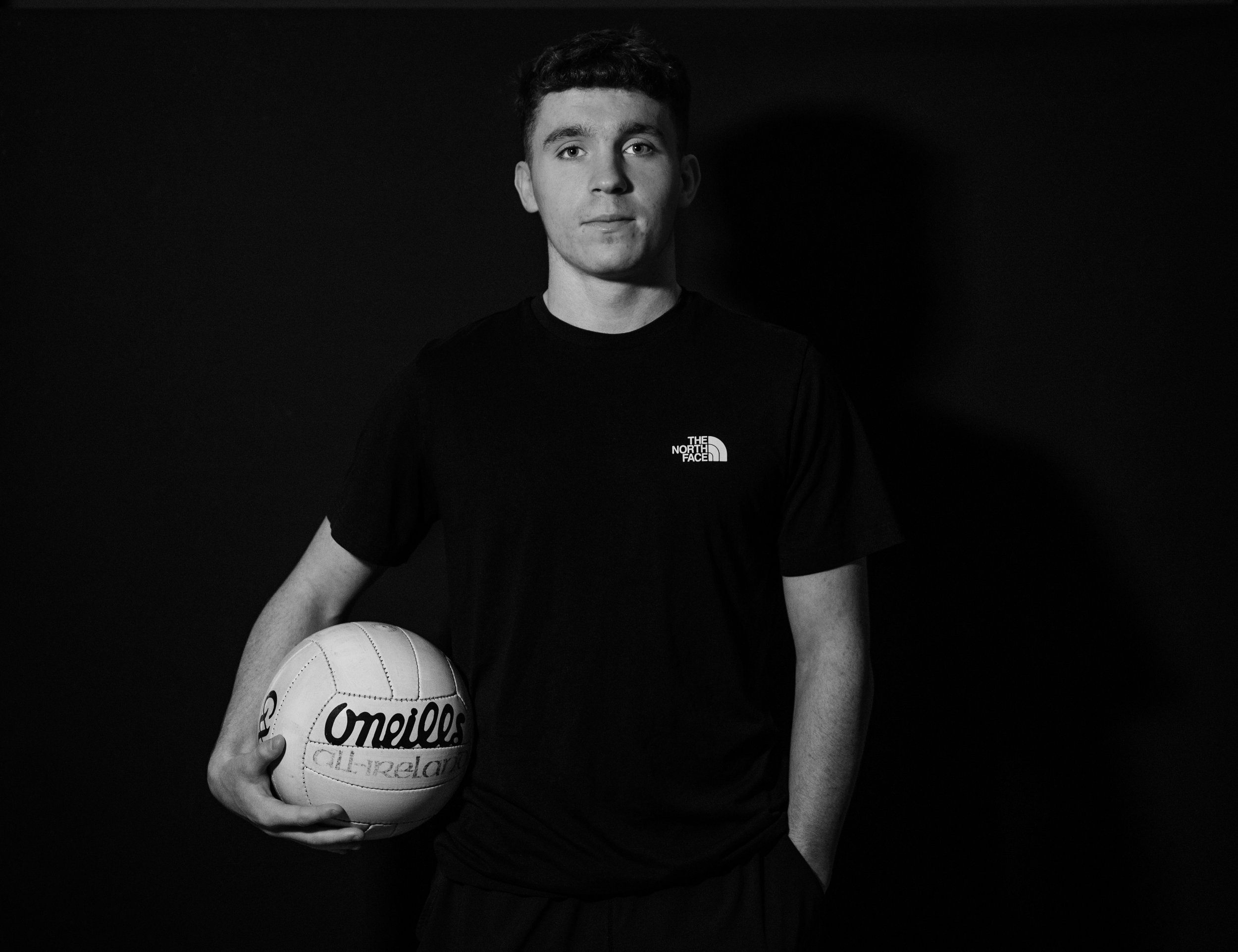 A young man holding a ball, wearing a black T-shirt, against a dark background.