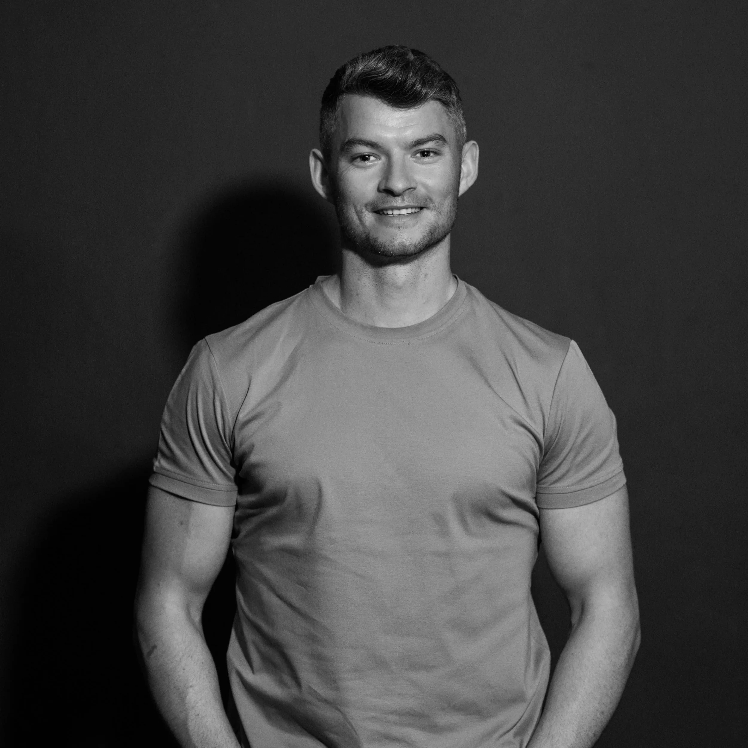 Black and white photo of a young man with short hair, wearing a fitted T-shirt, smiling, standing against a plain dark background.