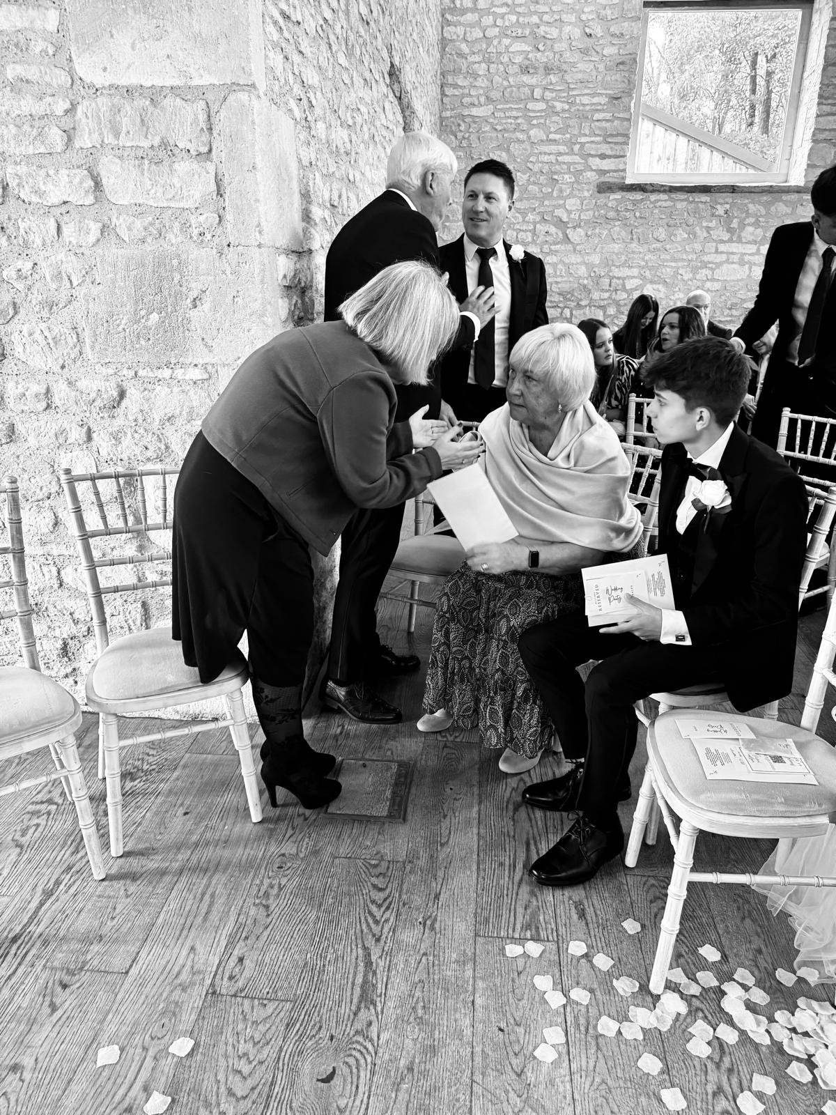 People dressed in formal attire, sitting and standing, interacting in a rustic indoor setting with stone walls and wooden floors. Chairs are arranged in rows with scattered flower petals on the floor.