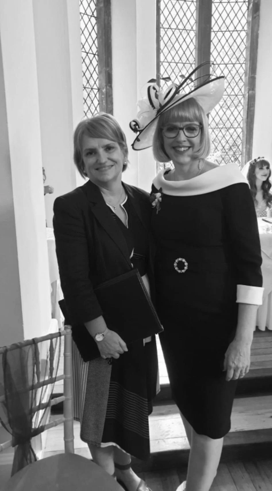 Two women standing together in formal attire, one wearing a decorative hat and glasses, indoors next to a window with a checkered grid pattern. They appear to be at a special event.