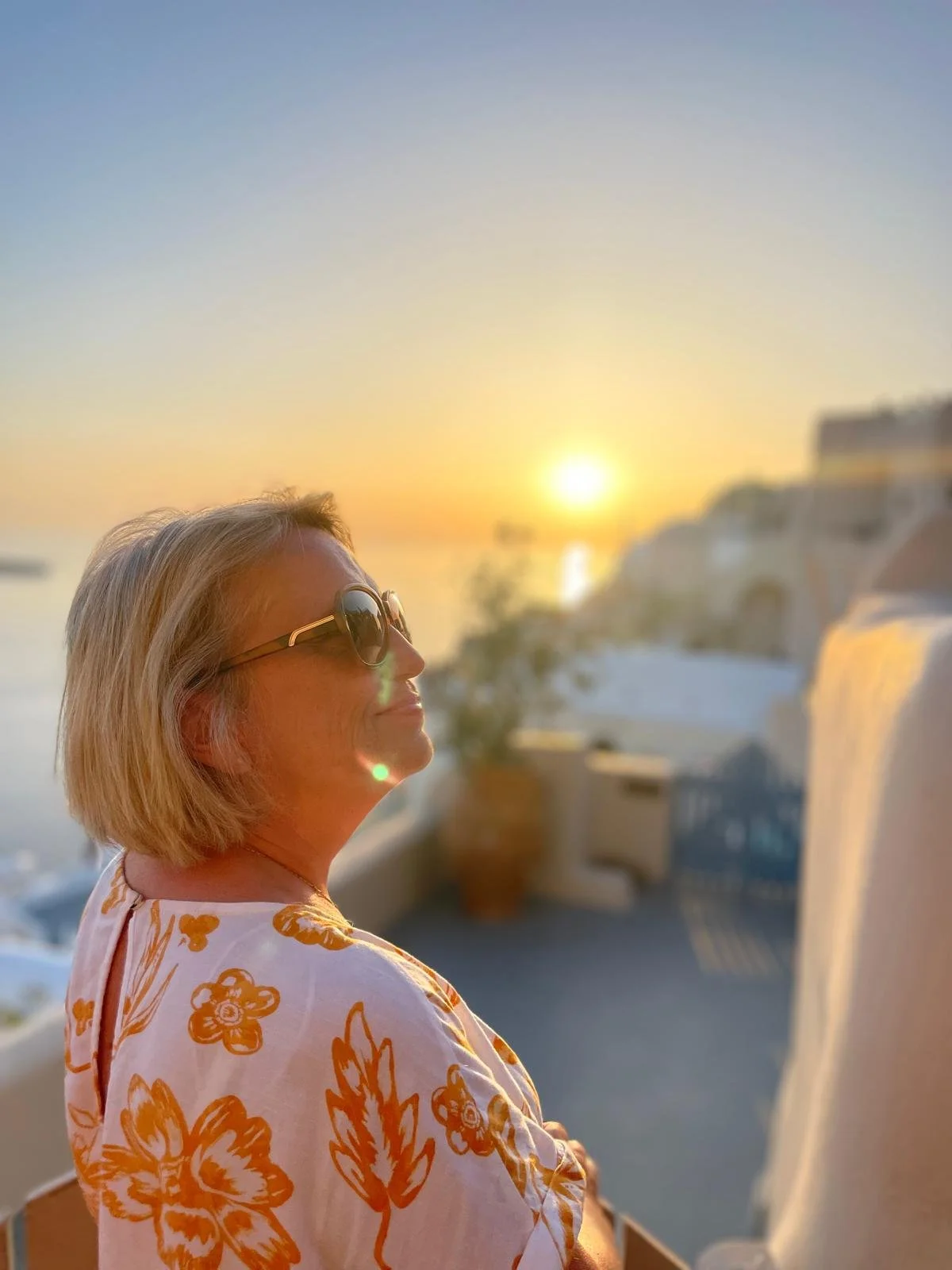 Person wearing sunglasses and a floral shirt enjoying a sunset view on a balcony.