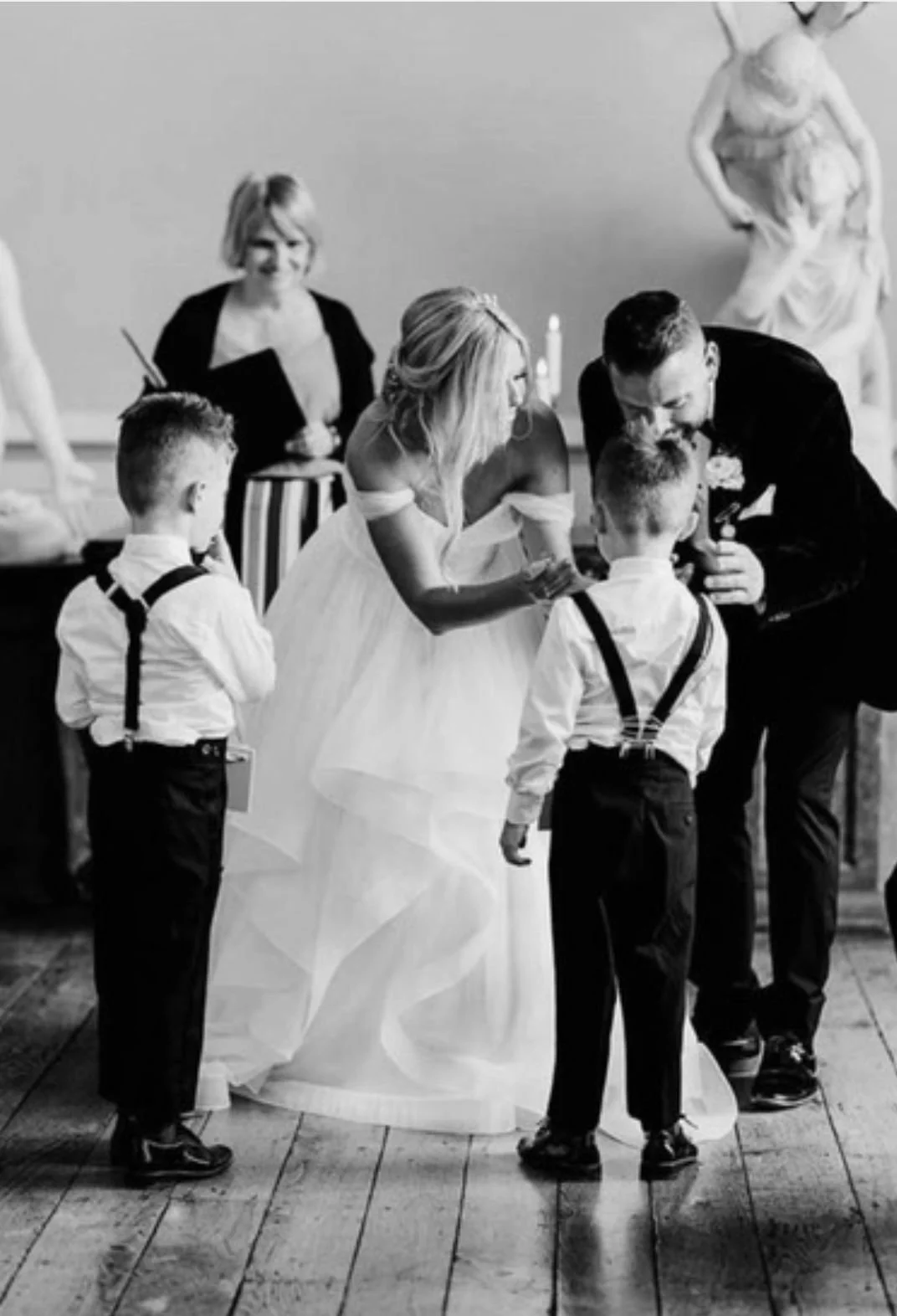 Bride and groom with young boys at a wedding ceremony, with an officiant and statues in the background.