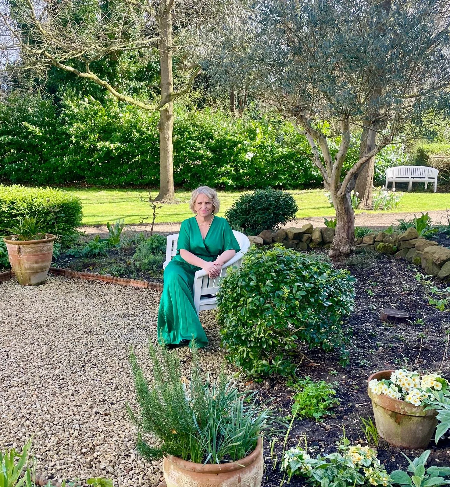 Woman in a green dress sitting on a white chair in a garden with trees and plants.