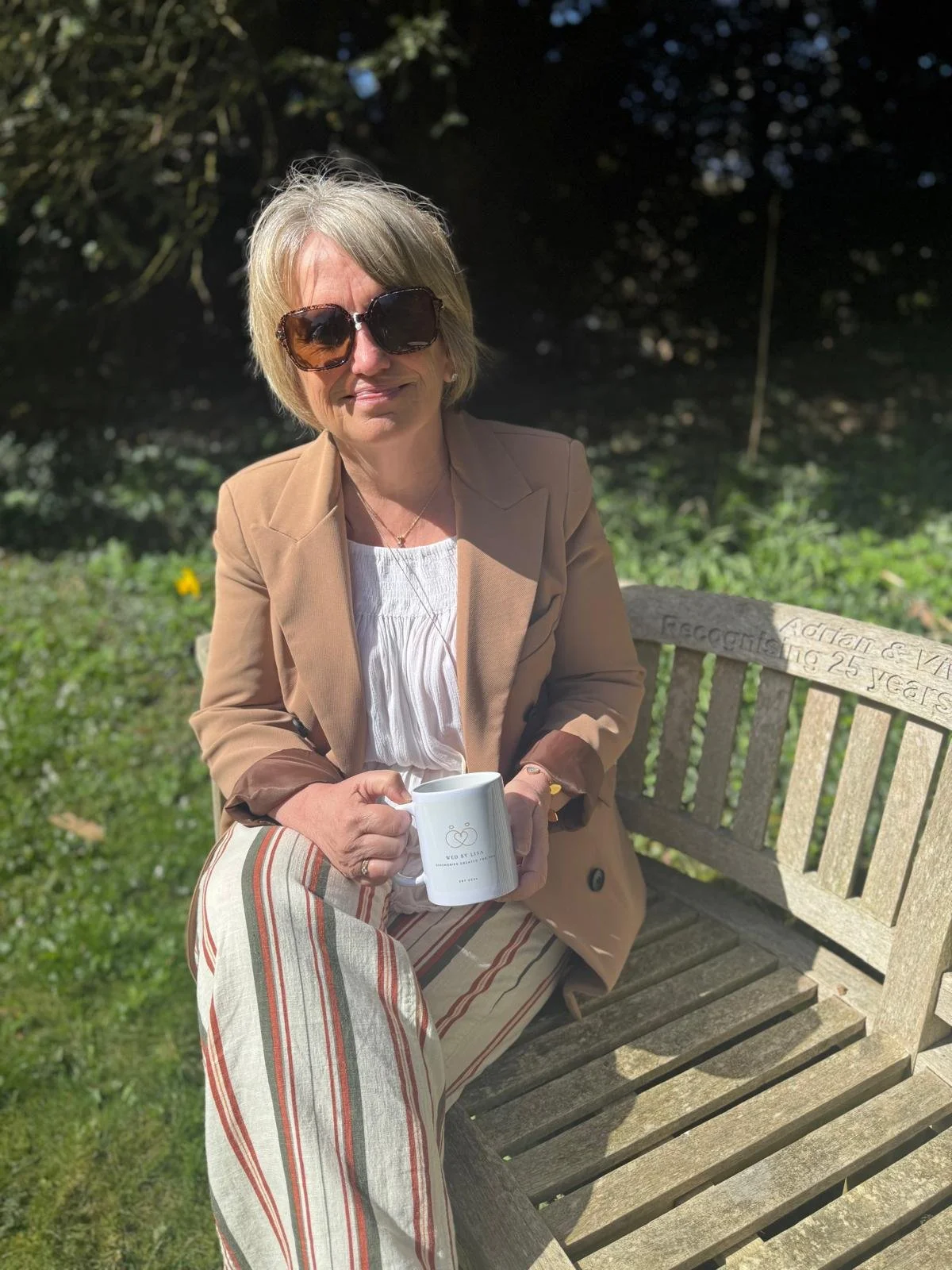 Woman with sunglasses sitting on a wooden bench holding a mug, wearing a brown blazer and striped pants, outdoors in a garden setting.