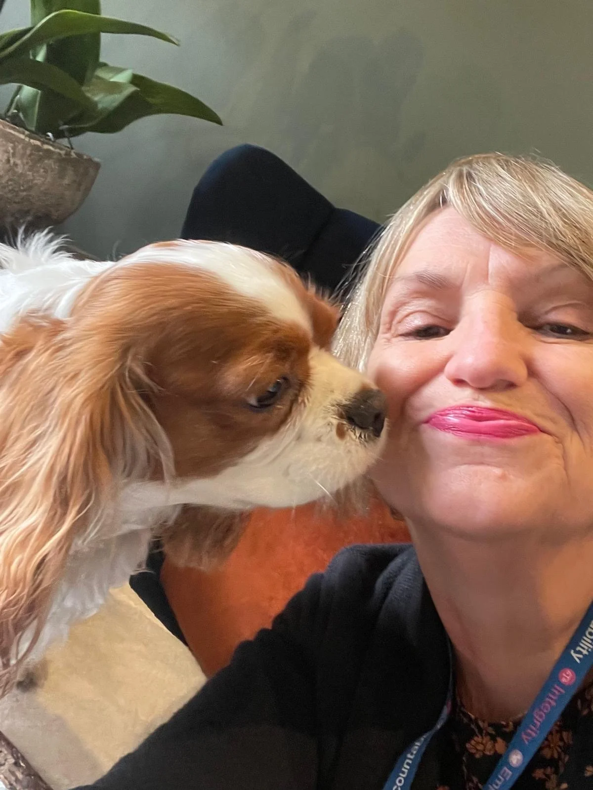 A woman smiling as a Cavalier King Charles Spaniel dog nuzzles her face. They are indoors with a green potted plant in the background.