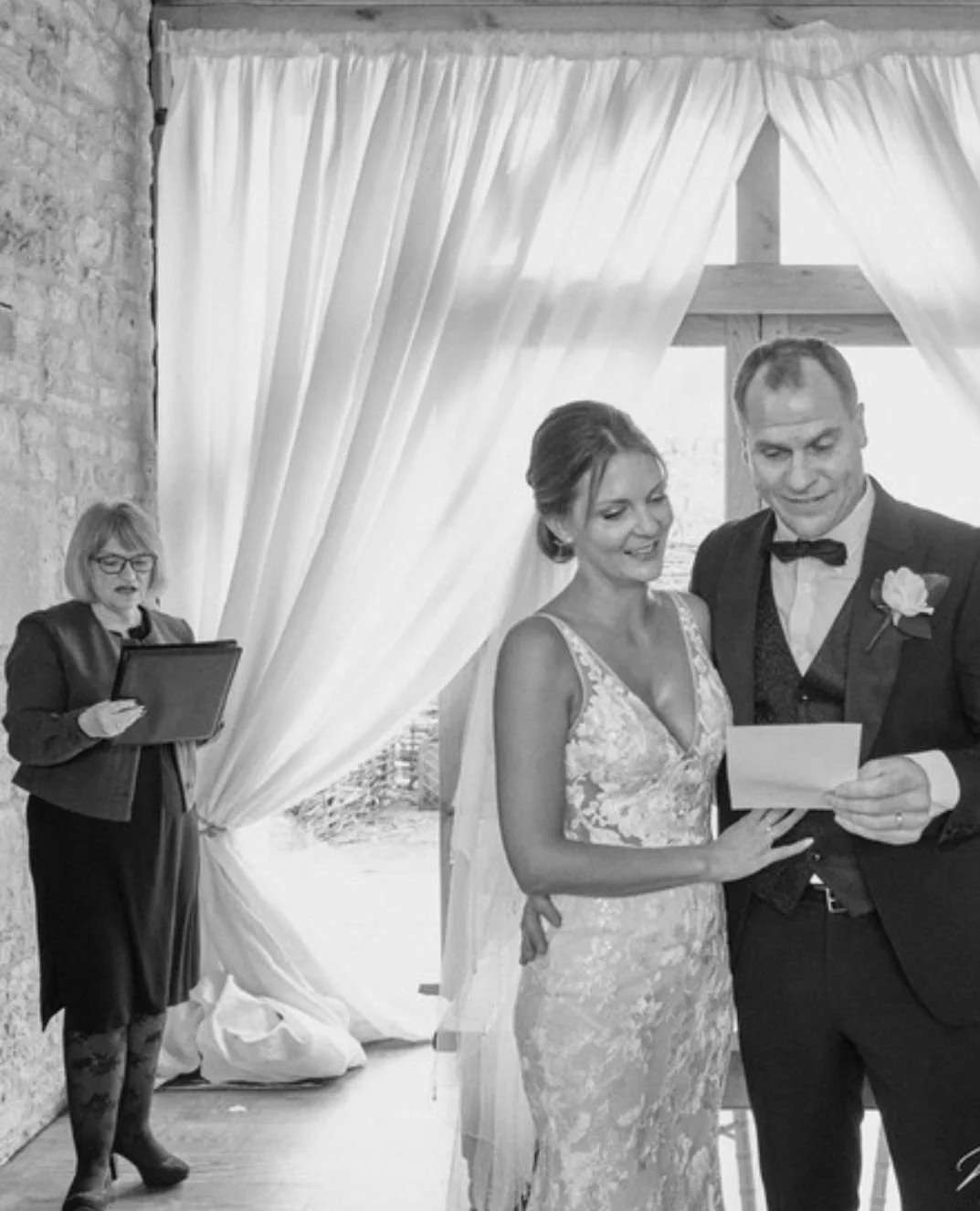 Bride and groom reading vows, officiant overseeing, draped curtains in background, wedding ceremony.