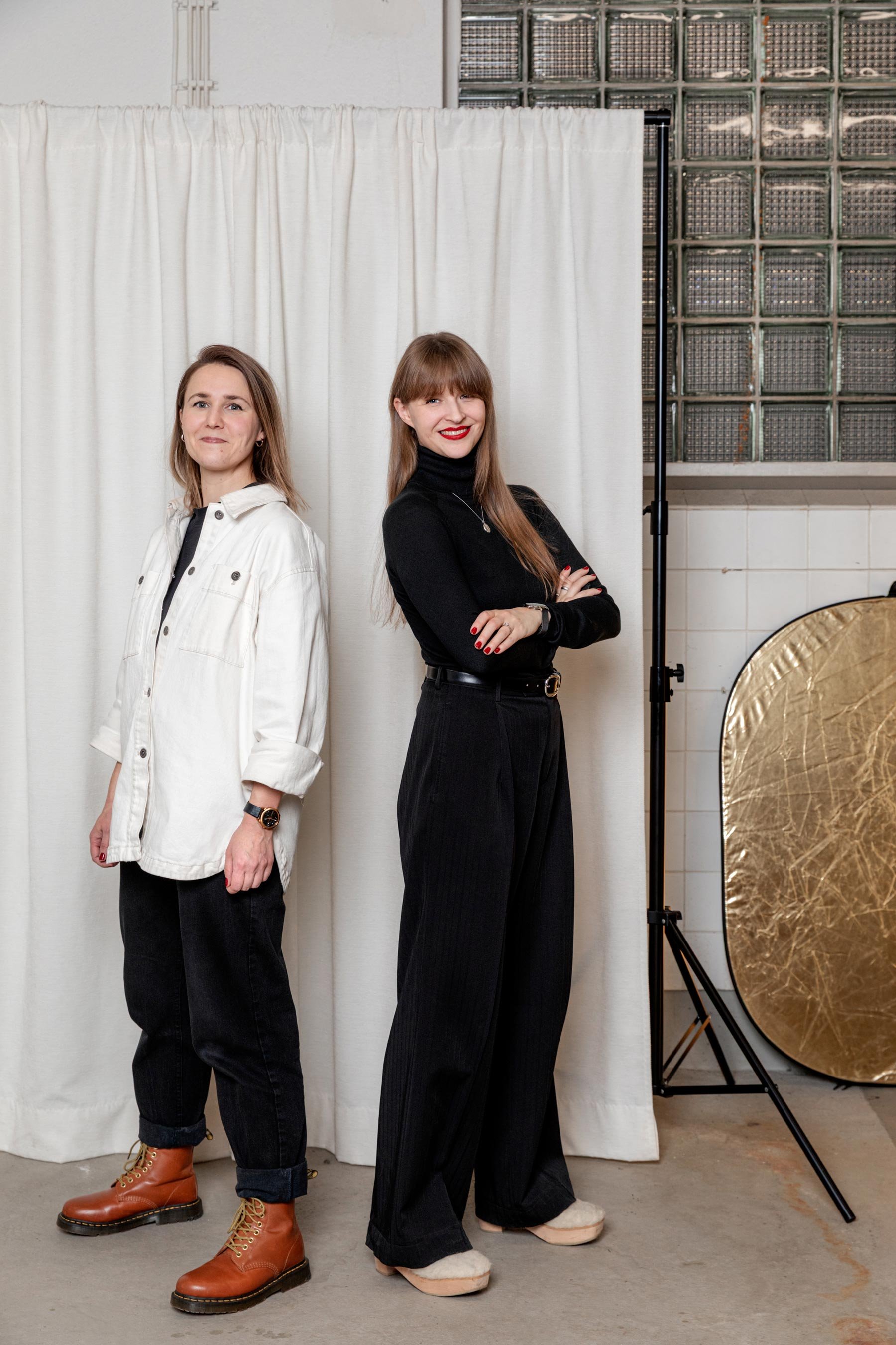 Anu ja Emmi katsovat kameraan ja hymyilevät taustallaan verho ja reflectori rekvisiittana