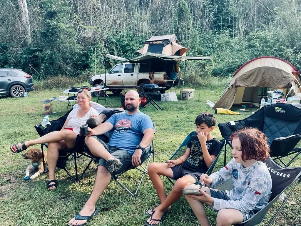 Family of four and their dog relaxing in camping chairs at a grassy campsite with a roof top tent and ground tent in the background.