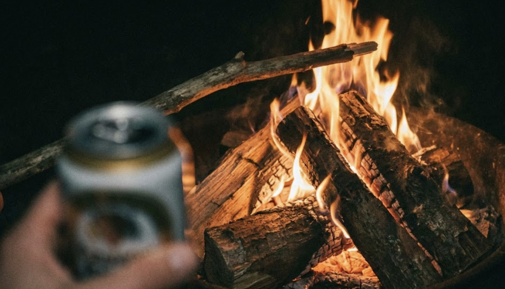 Close-up of a rustic campfire at night featuring a simple cooking grate and a hand holding a beer, representing the minimalist approach to open fire cooking.