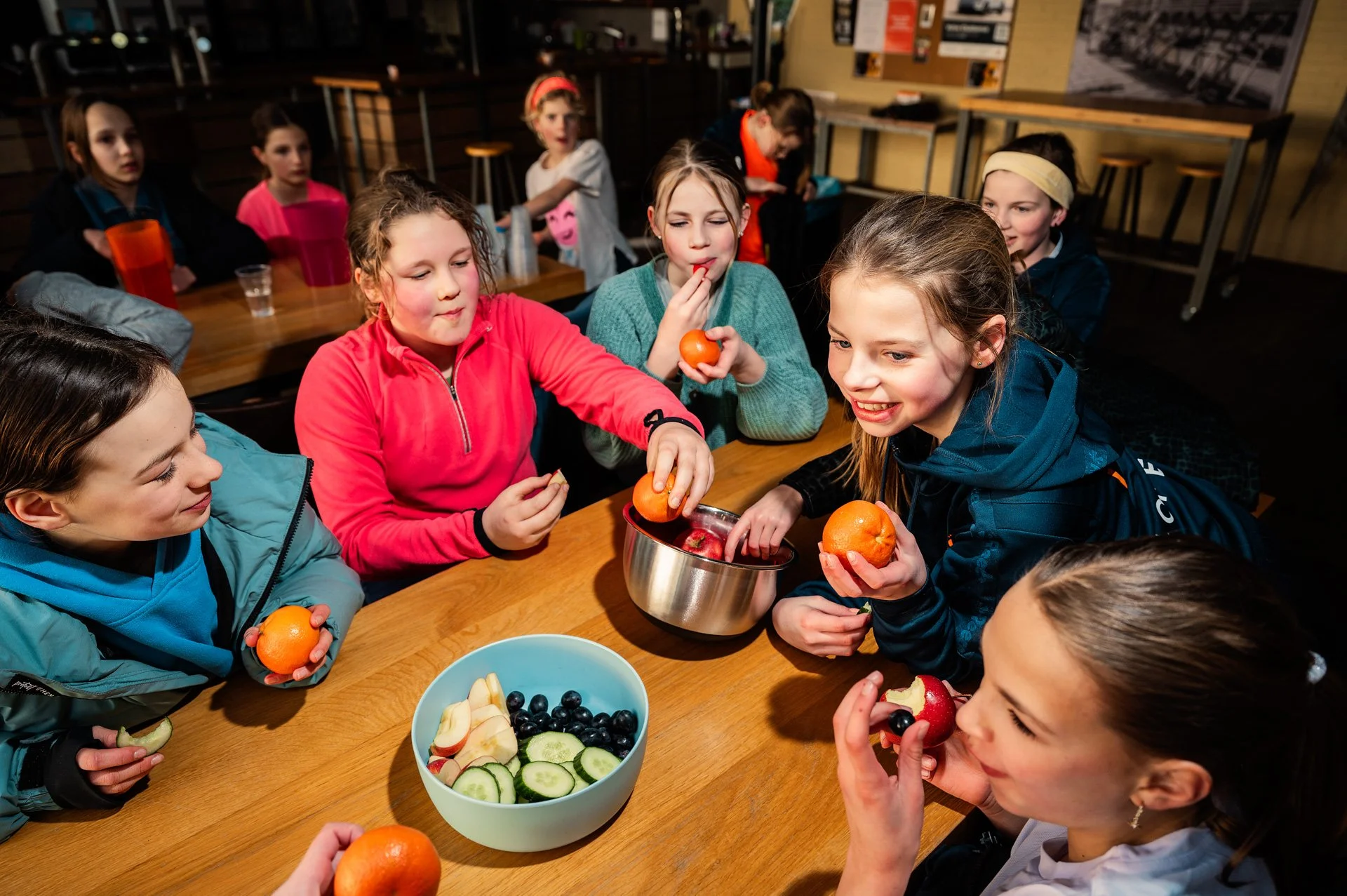 Verschillende kinderen zitten rond een tafel met fruit, zoals appels, sinaasappels, druiven en komkommers, en genieten van het eten tijdens een feestje of groepsactiviteit.