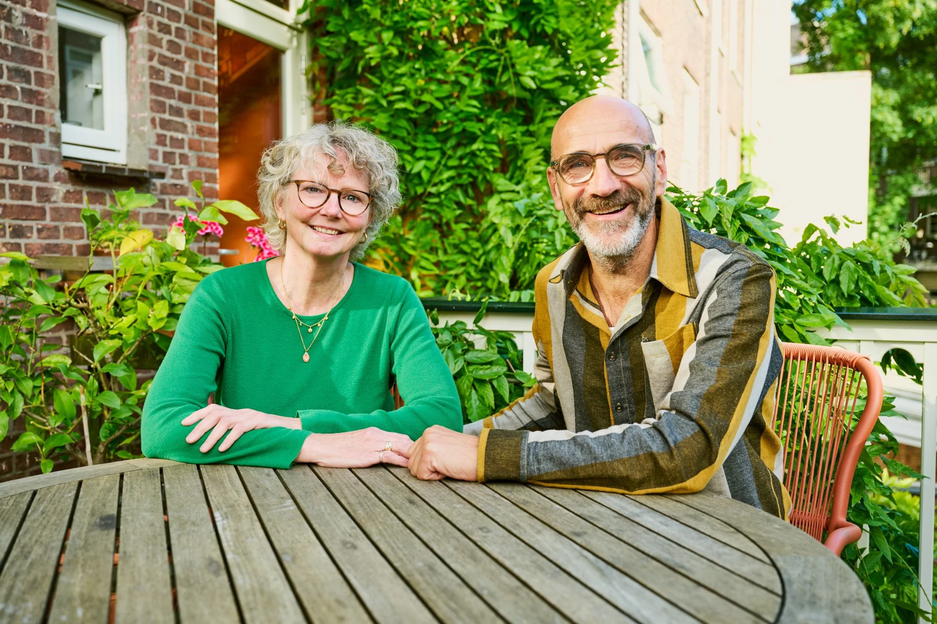 Een echtpaar zit aan een tuintafel en lacht vriendelijk in de camera.