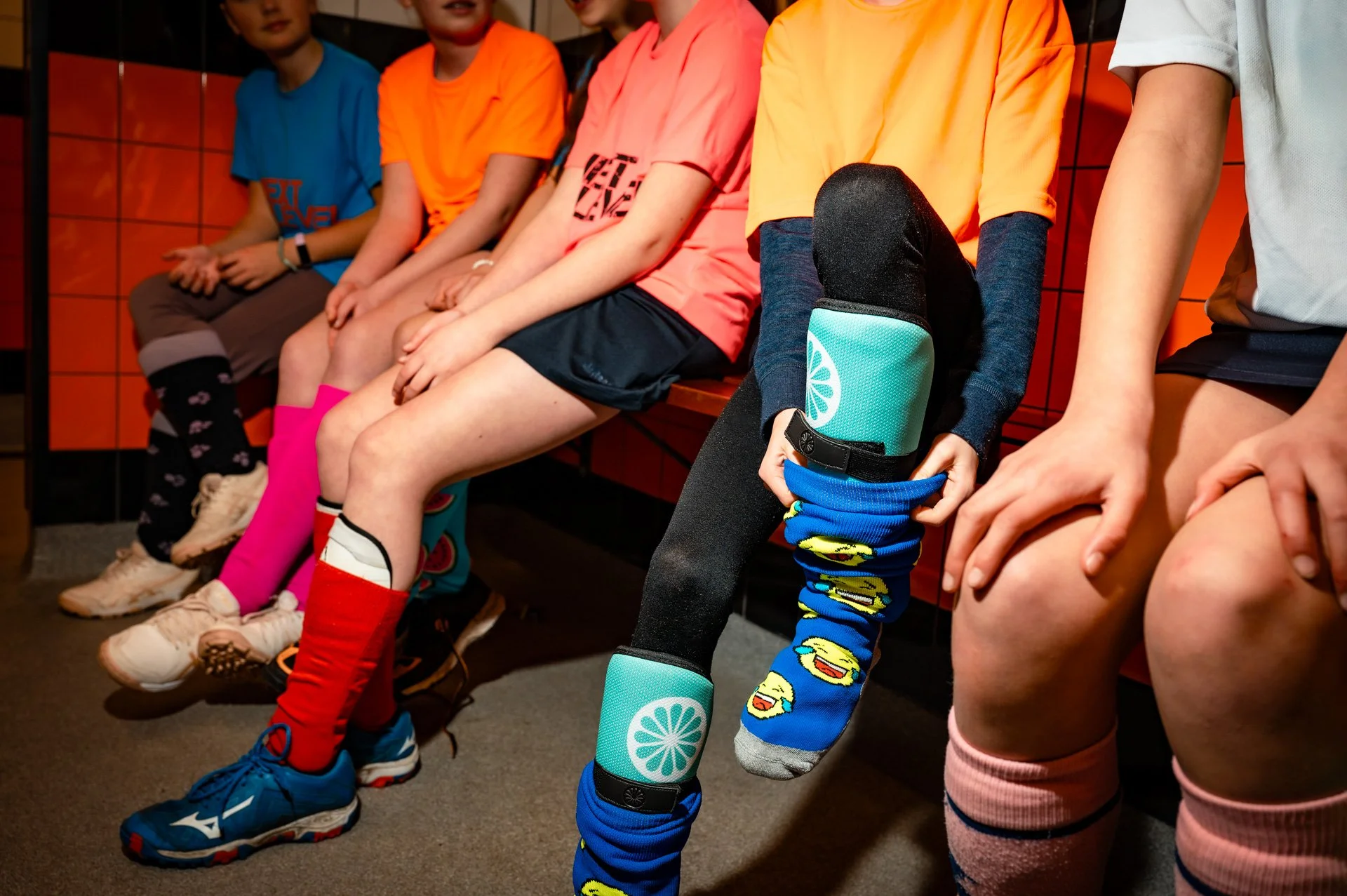 Vijf kinderen zitten op een bank met hun handen op hun knieën, gekleed in kleurrijke sportkleding en sokken, in een kleedkamer of kleedruimte.