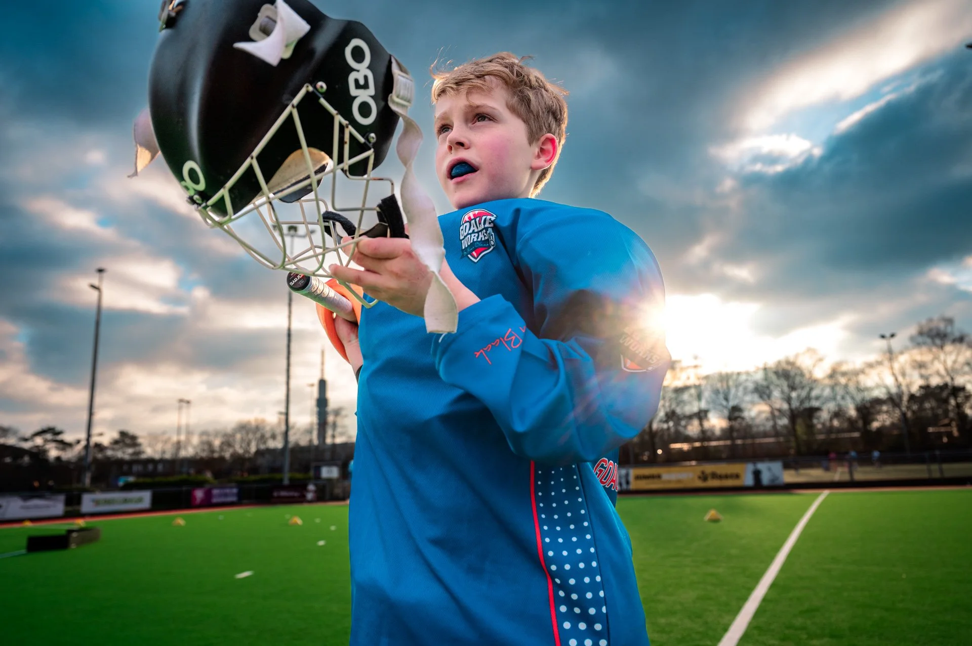 Een jonge hockeyspeler in een blauwe sportoutfit met een stick en helm op een hockeyveld, onder een bewolkte hemel met de zon die schijnt.