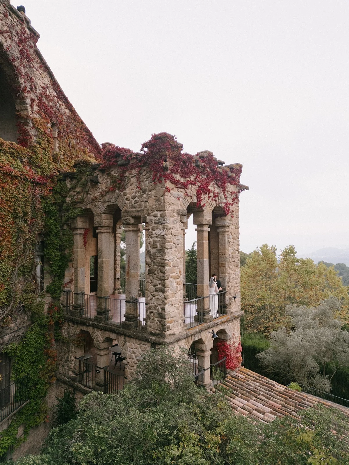 Couple standing on the balcony of a stone tower covered in red autumn ivy at a Catalan wedding venue