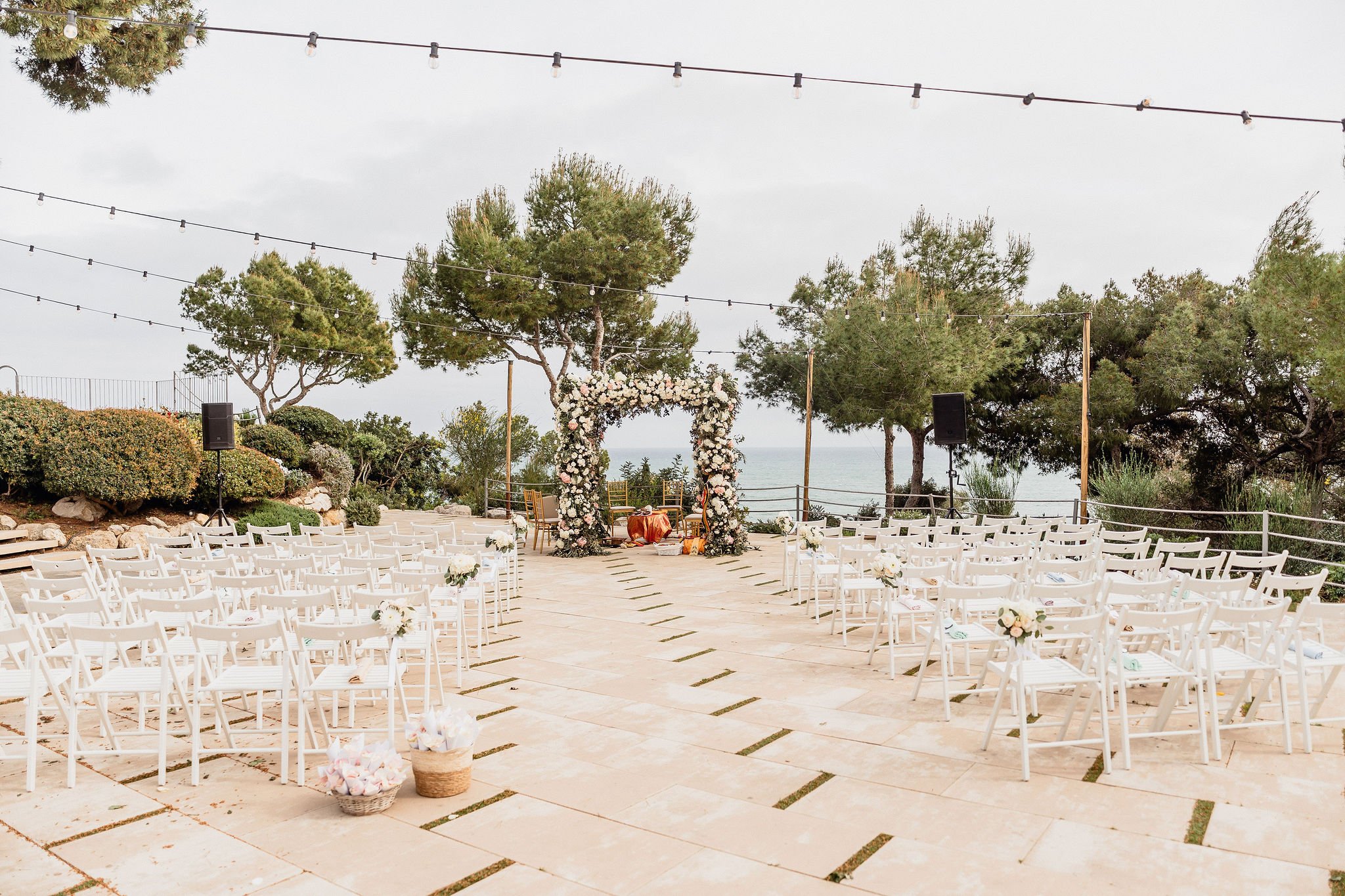 Outdoor ceremony set-up with white chairs, a floral arch and string lights by the Mediterranean Sea in April