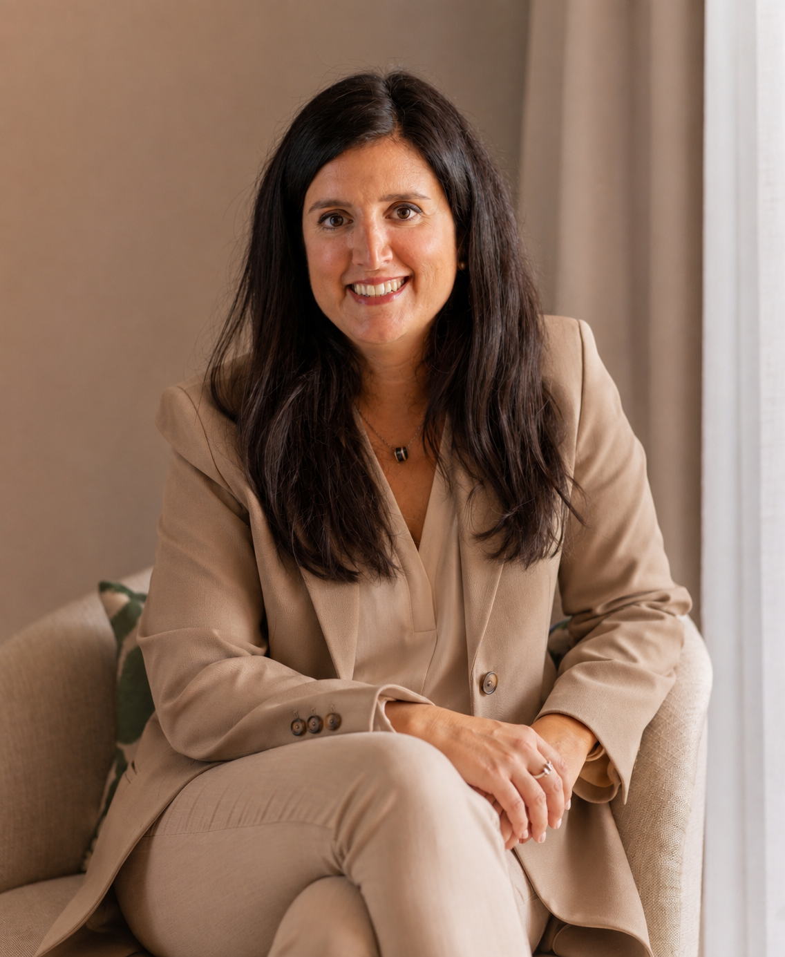 A woman with long dark hair wearing a beige suit, sitting on a beige chair in a room with neutral-colored walls and a window with curtains, smiling at the camera.