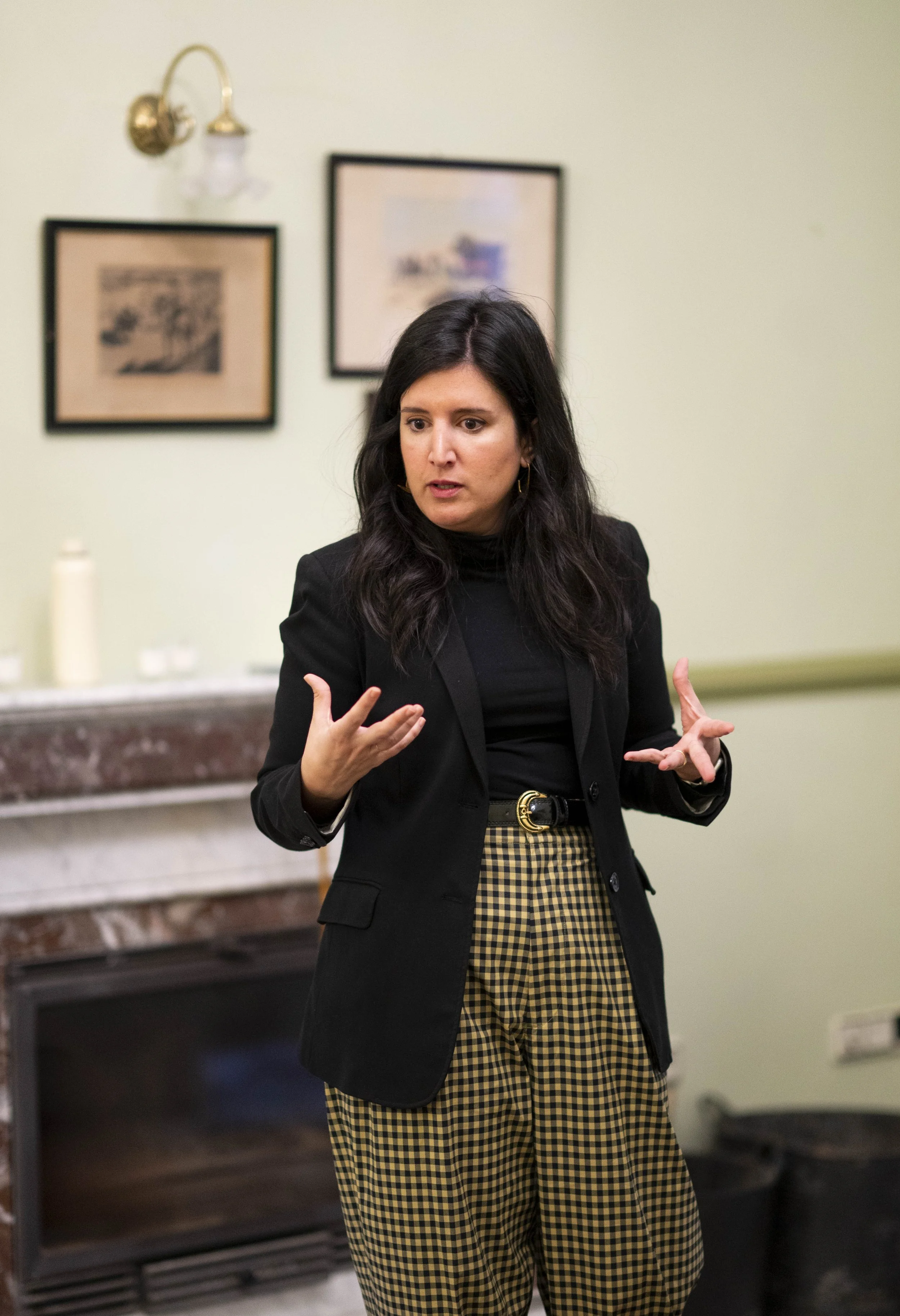 A woman with dark hair wearing a black blazer and yellow plaid pants, speaking and gesturing with both hands in a room with green walls, two framed pictures, and a fireplace.