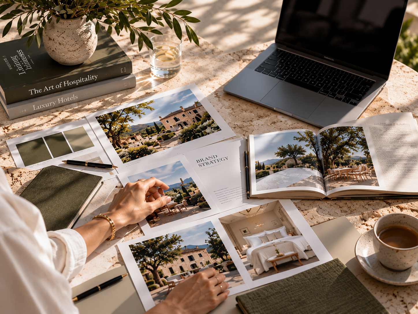A person working at an outdoor desk with travel planning materials, including brochures, a laptop, books, a notebook, and a cup of coffee, surrounded by shadows and sunlight.