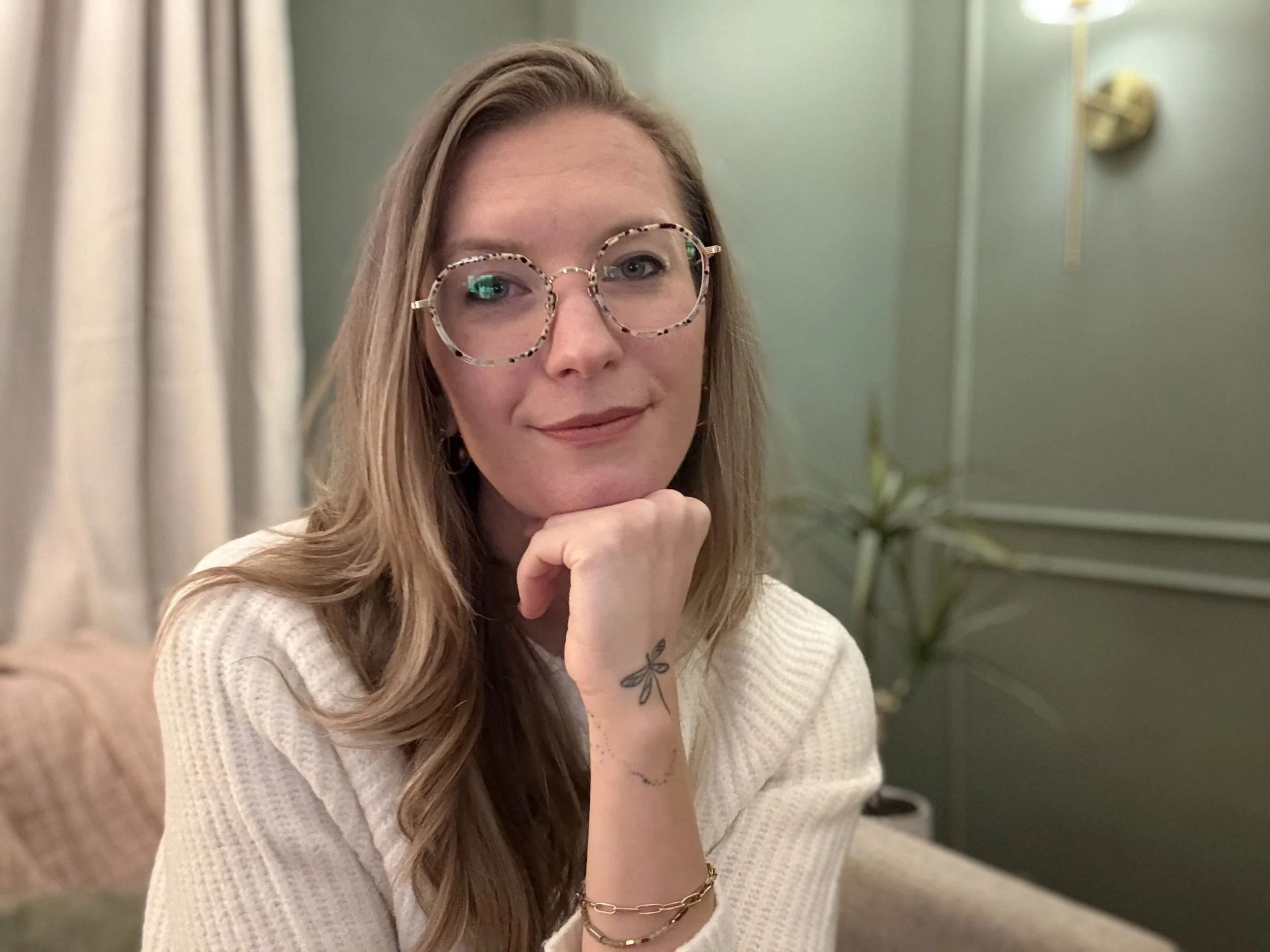 A woman with long blonde hair, glasses with tortoiseshell frames, and a white sweater sitting indoors with a hand under her chin, smiling gently, in a room with light green walls, beige curtains, a potted plant, and a wall-mounted light fixture.