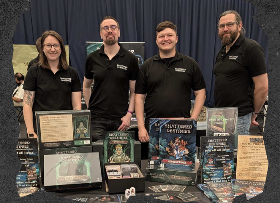Four people standing behind a table at a booth selling tabletop game products, with various game boxes, promotional materials, and cards displayed on the table, all wearing matching black polo shirts with a logo.