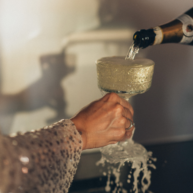 A bride's hand holding out a glass of champagne which is being topped up to the point of overflowing