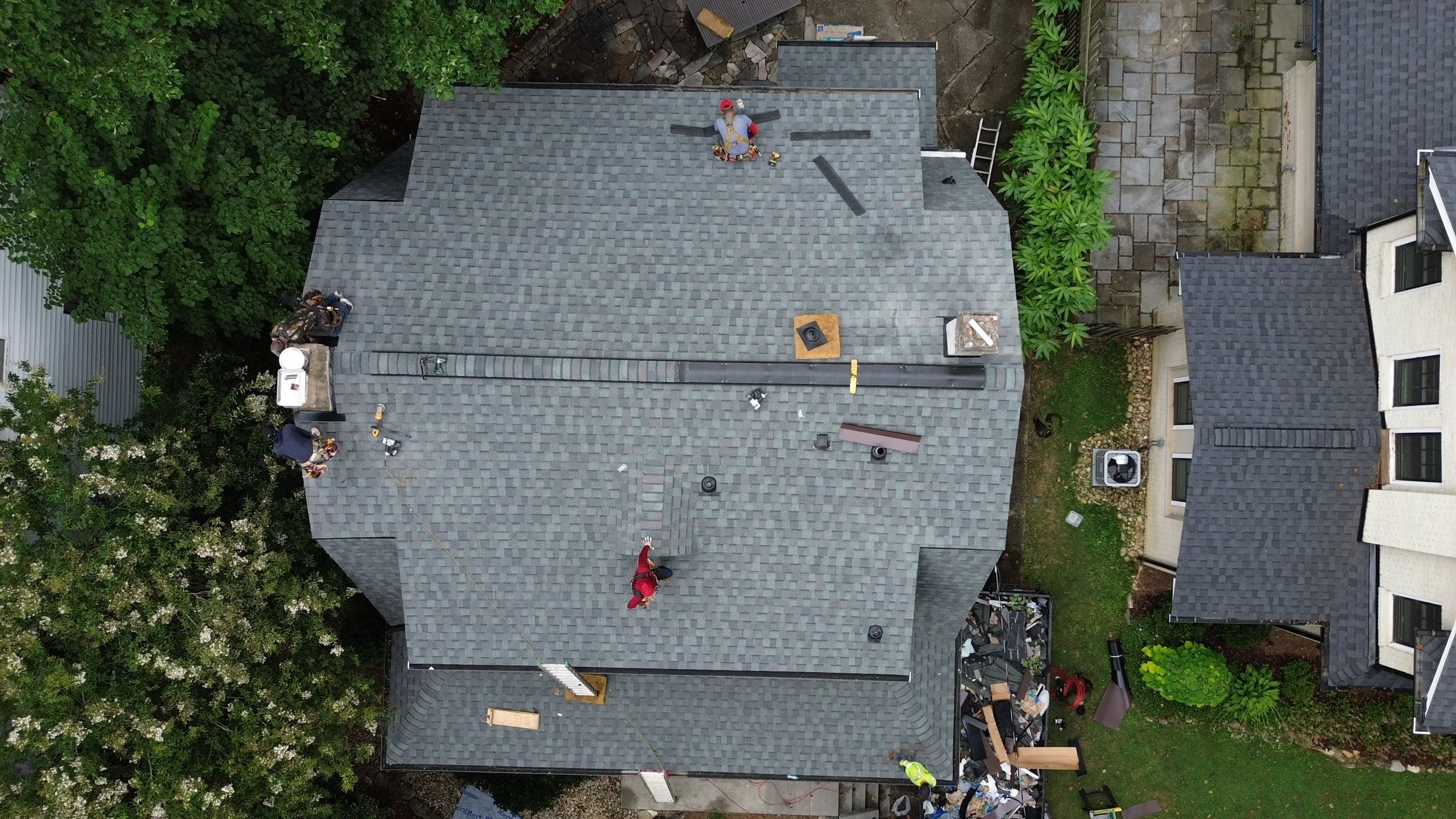 Aerial view of a house roof with gray shingles, surrounded by green trees and a cloudy sky.