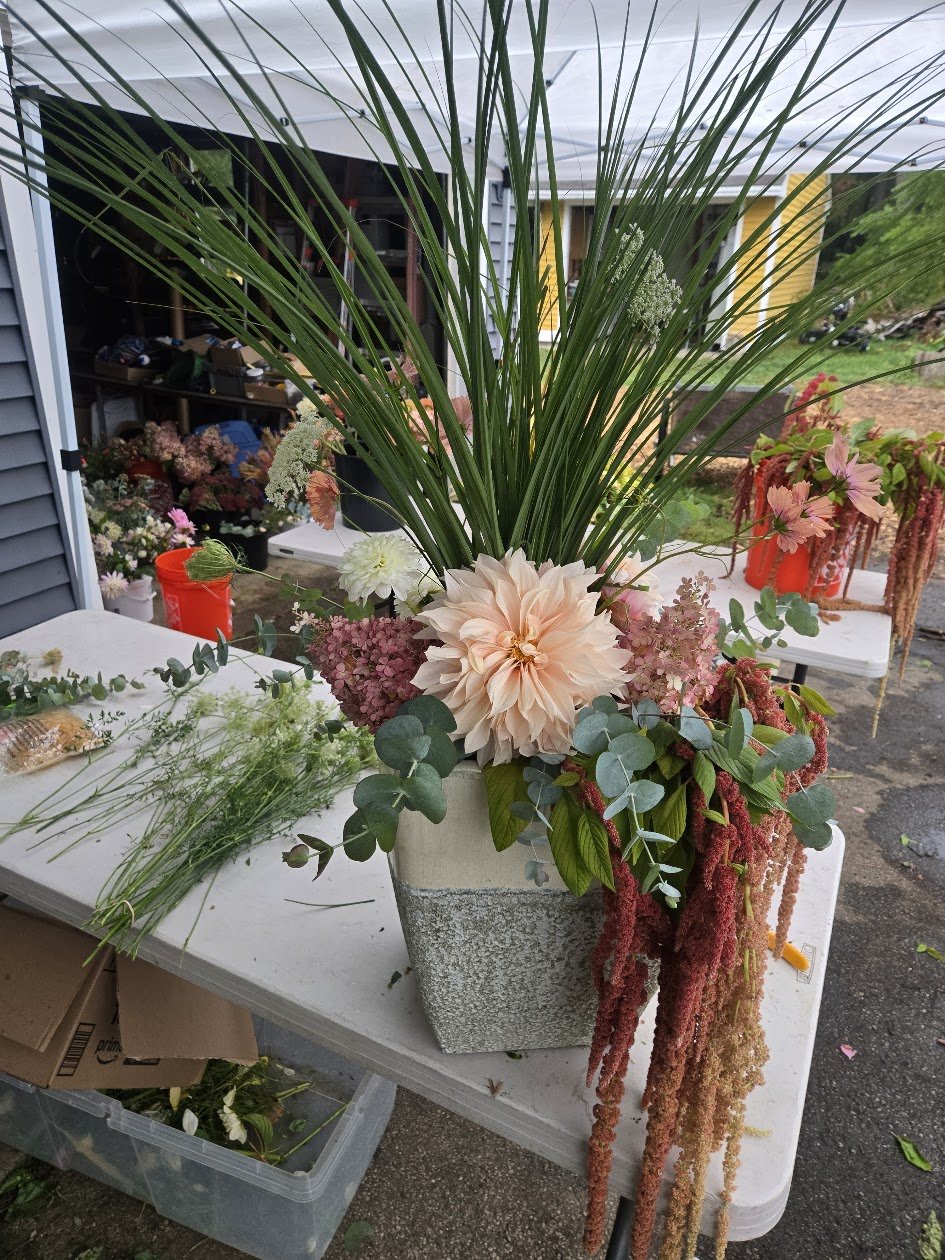 A large floral arrangement in a rectangular concrete vase featuring tall green grass, pink dahlias, pink and burgundy amaranthus, and eucalyptus leaves, set on a white table outdoors.