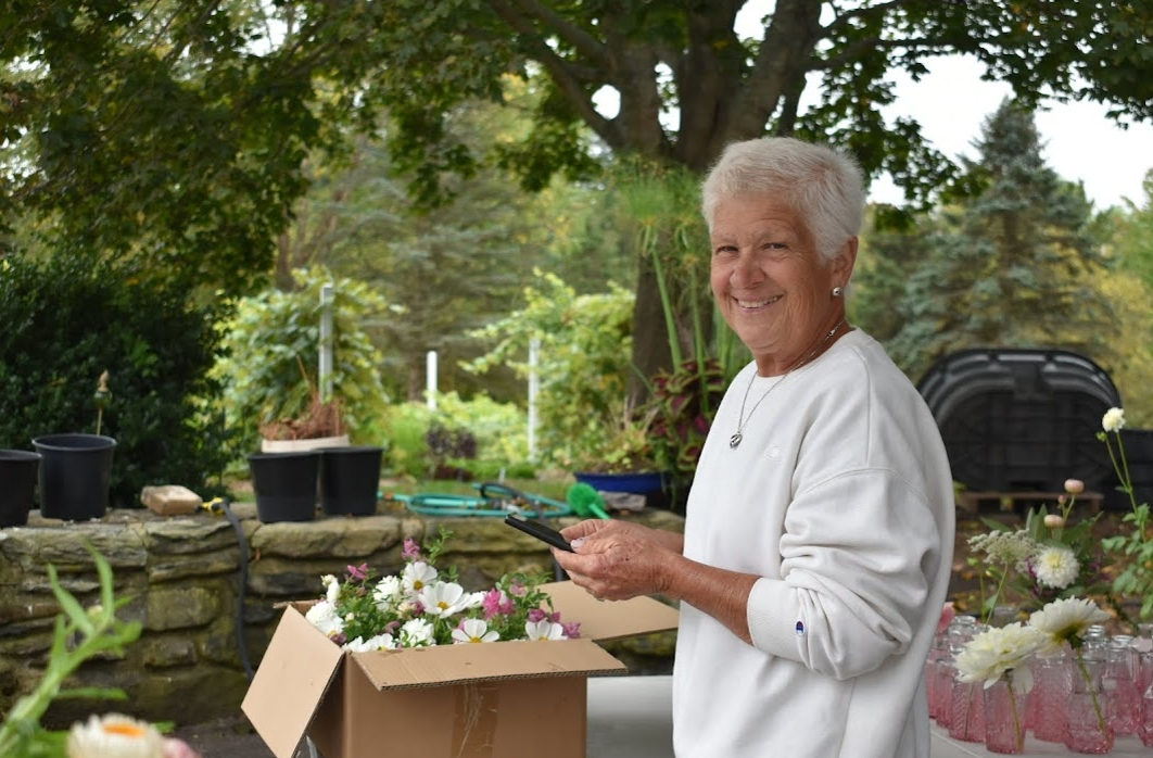 A smiling elderly woman standing outdoors in a garden, holding a cell phone, with a box of flowers and gardening supplies around her.