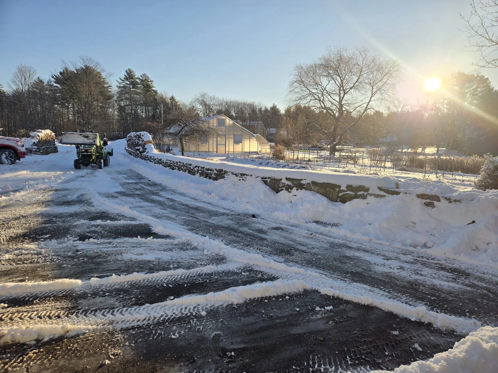 Snow-covered driveway with tire tracks and a tractor clearing snow, along with a red vehicle, in a rural area with trees, a greenhouse, and a stone wall, under a bright winter sun.