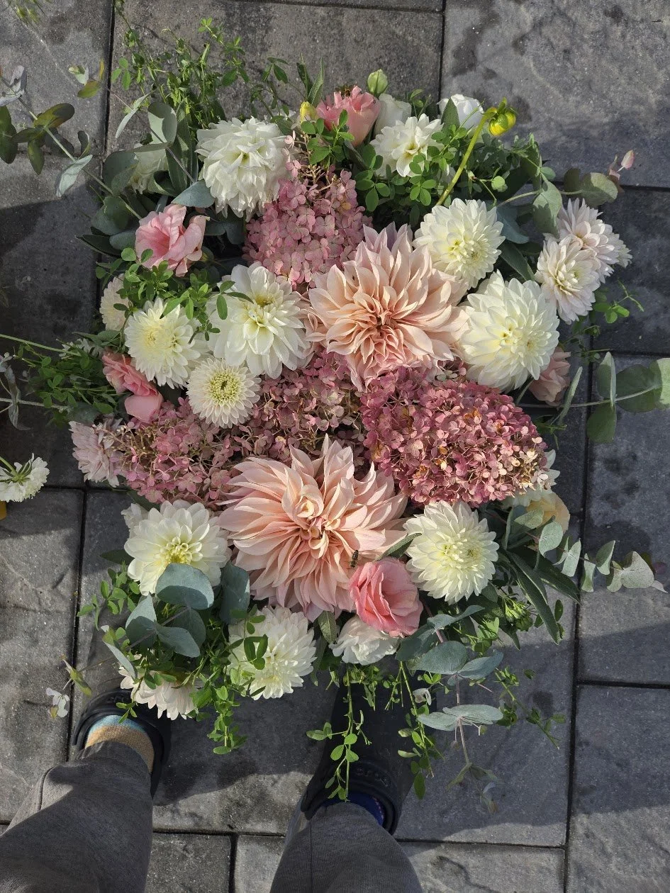 A top-down view of a bouquet of mixed flowers, including dahlias, roses, hydrangeas, and greenery, placed on a stone pavement.