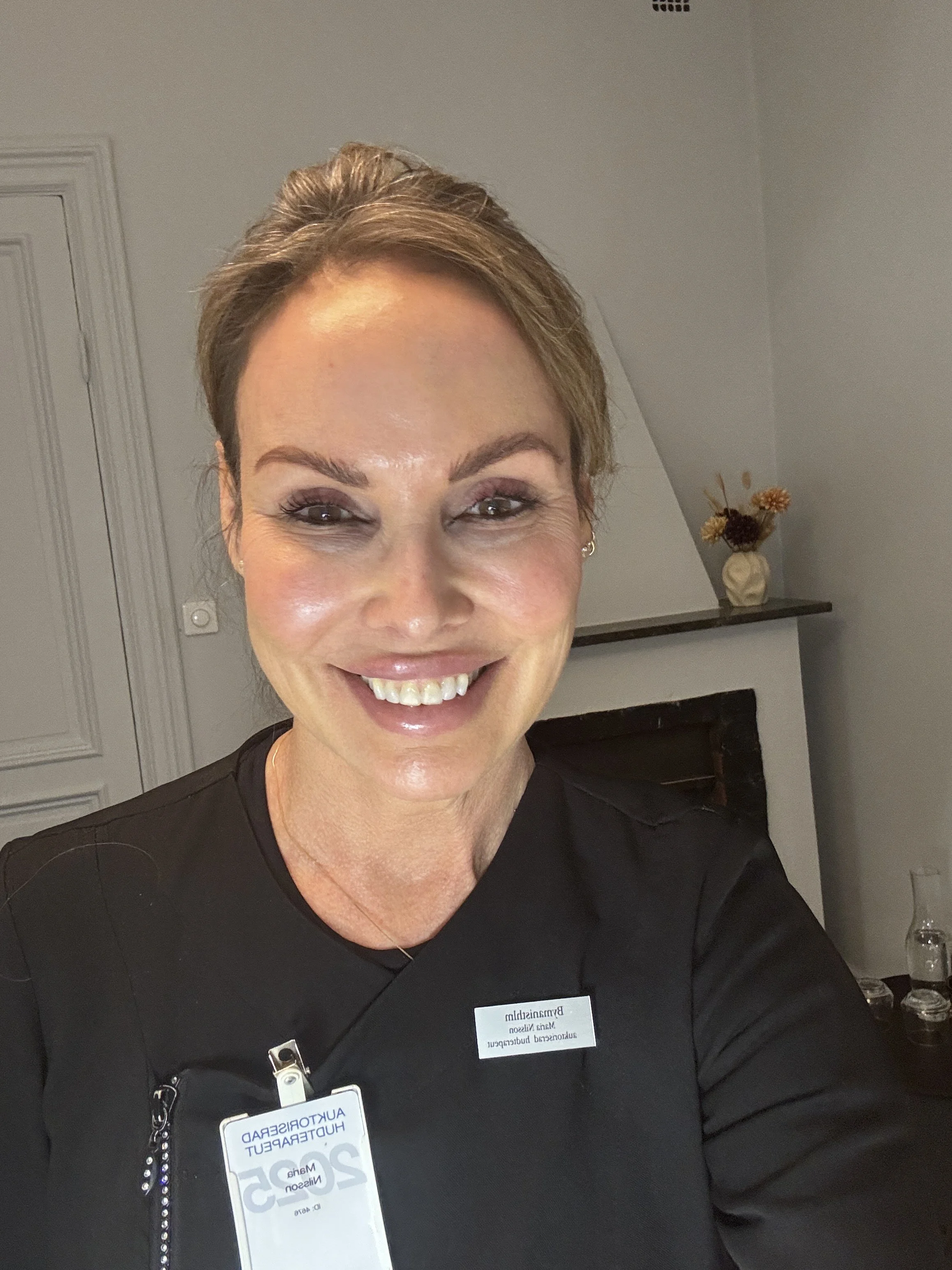 En kvinna med kort, ljusbrunt hår som tar en selfie inomhus. Hon bär svarta arbetskläder och har en namnskylt. Bakgrund med vit vägg, öppen spis, ett brunt hyllskåp med en vas och blommor, samt några glasflaskor på ett bord.