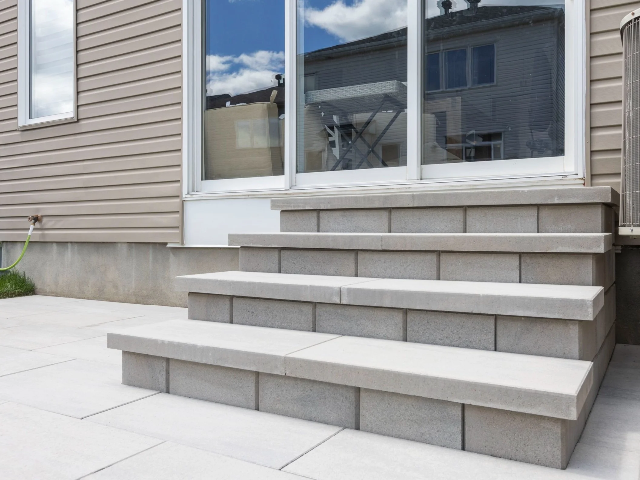 Image of newly installed concrete steps with a smooth surface and textured sides, leading up to a patio door with a reflection of the sky and another house.
