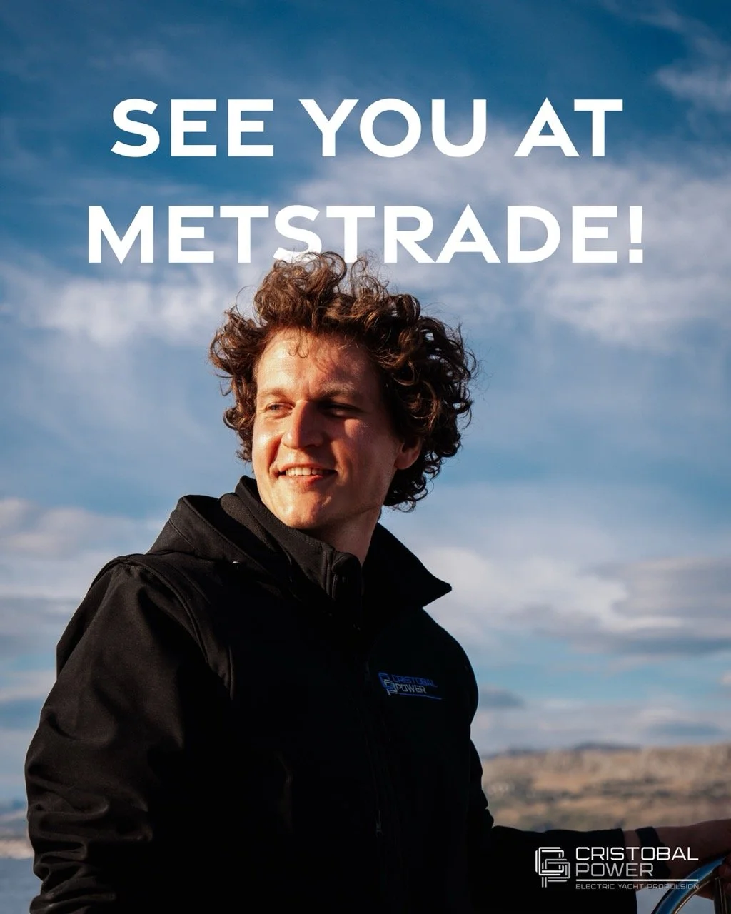 A man with curly hair wearing a black jacket with a Cristobal Power logo, smiling outdoors against a cloudy sky with a landscape in the background, with text saying 'See you at Metstrade!'