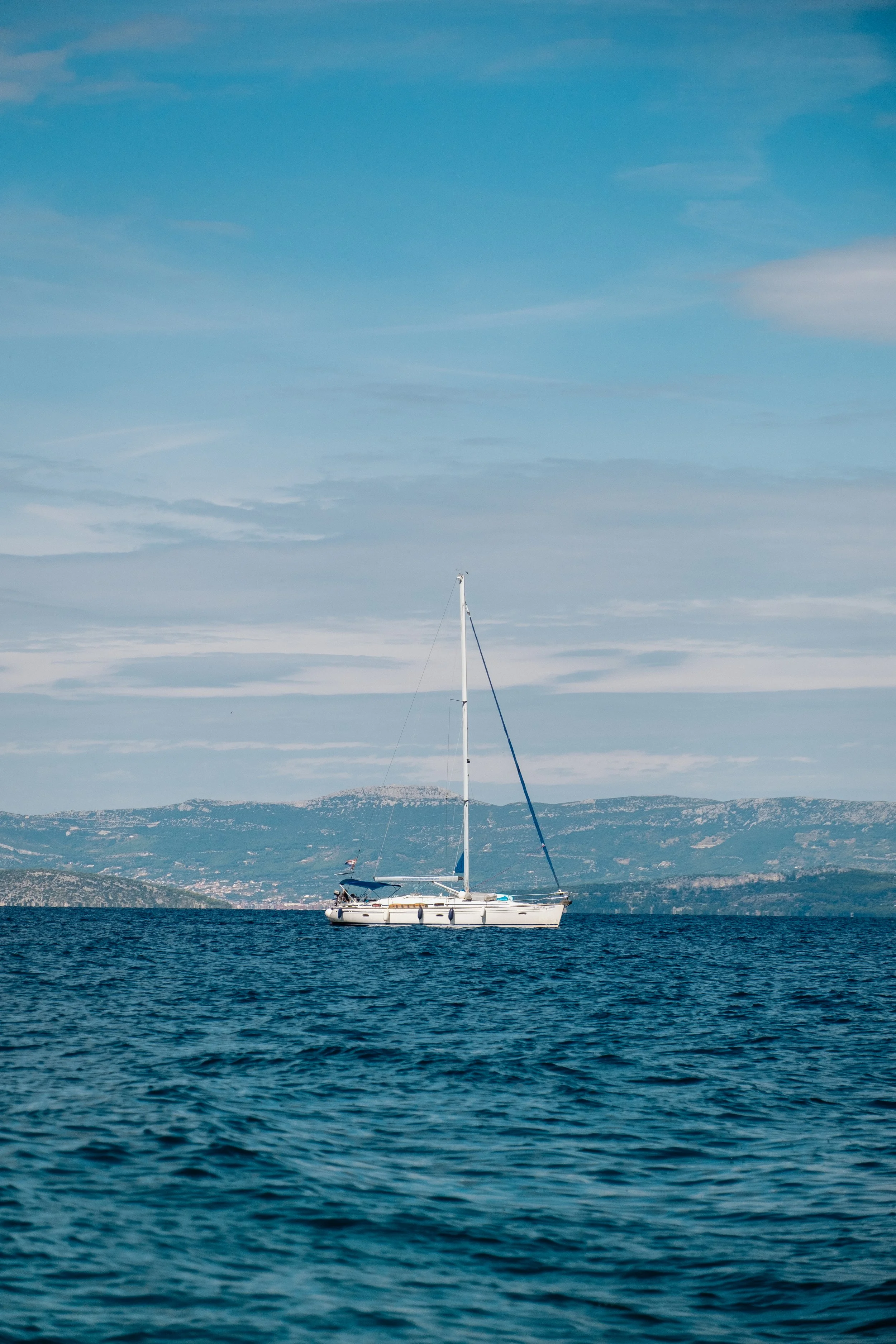 A sailboat floating on calm blue water with a mountainous landscape in the background under a partly cloudy sky.