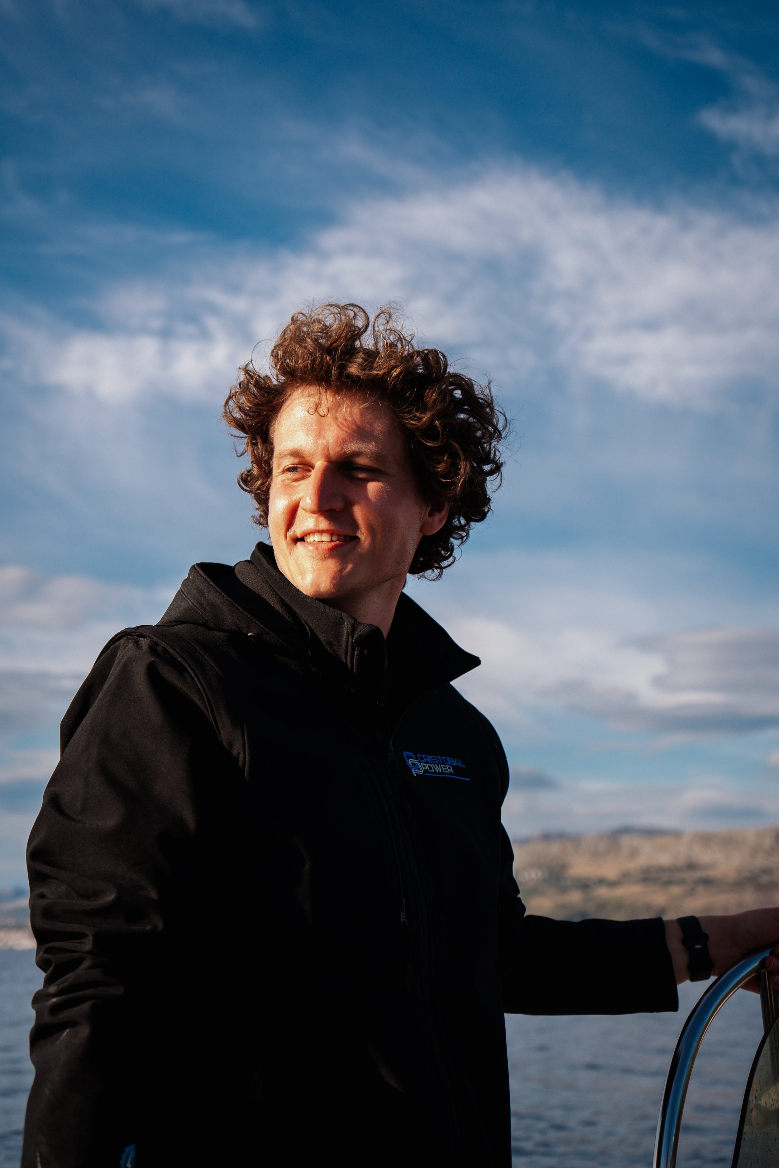 A man with curly hair smiling outdoors, wearing a black jacket, with a body of water and cloudy sky in the background.