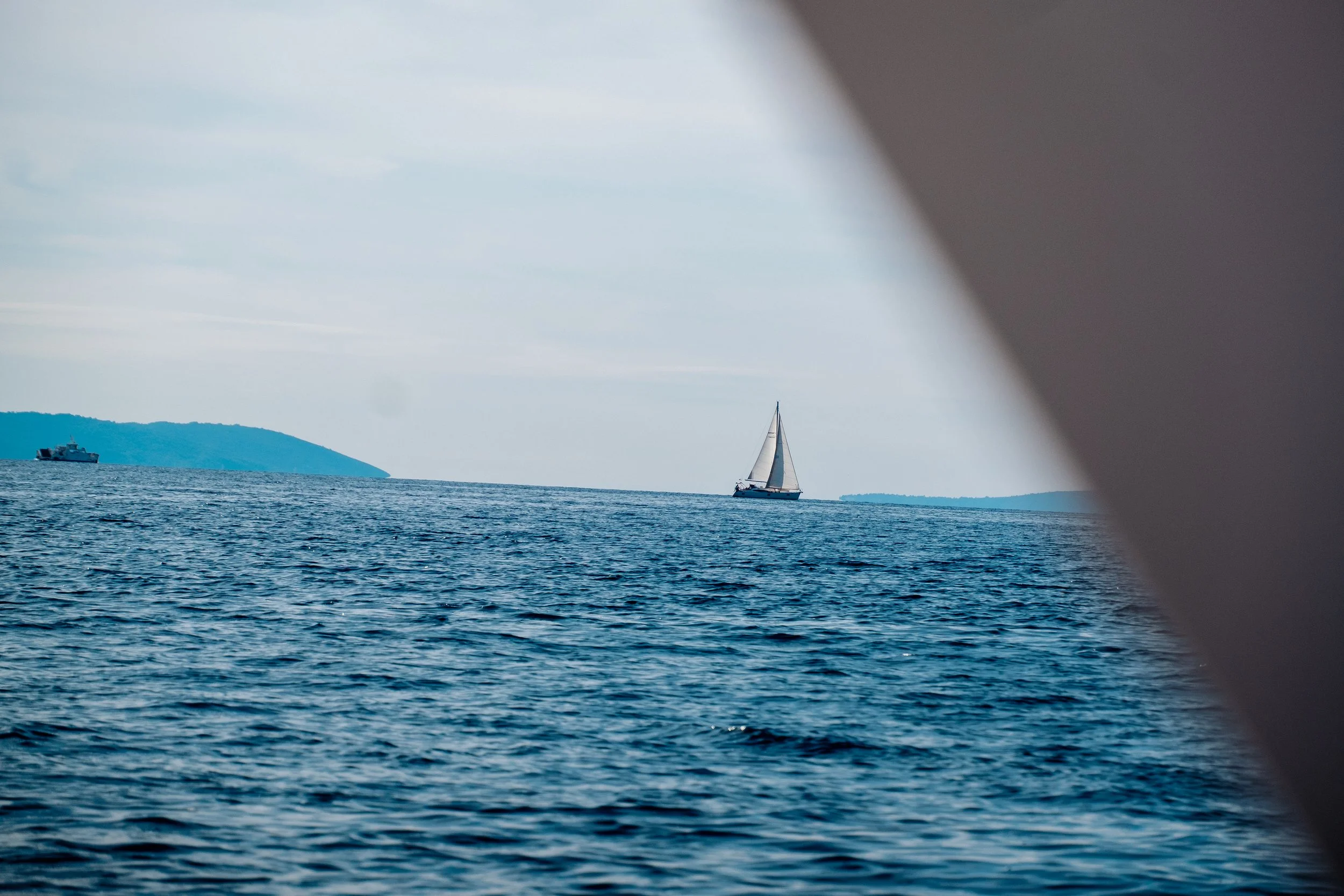 View of the ocean with a sailboat in the distance, flanked by two landmasses and partially obscured by a boat's edge.